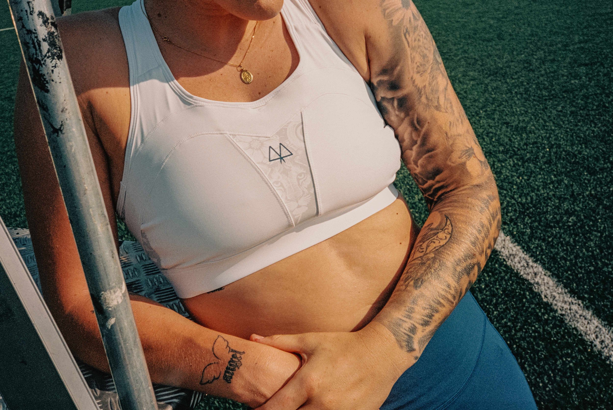 Close-up of a woman in workout attire sitting on a sports field, showing her muscular arms and tattoos, with a focus on her torso and arm tattoos.