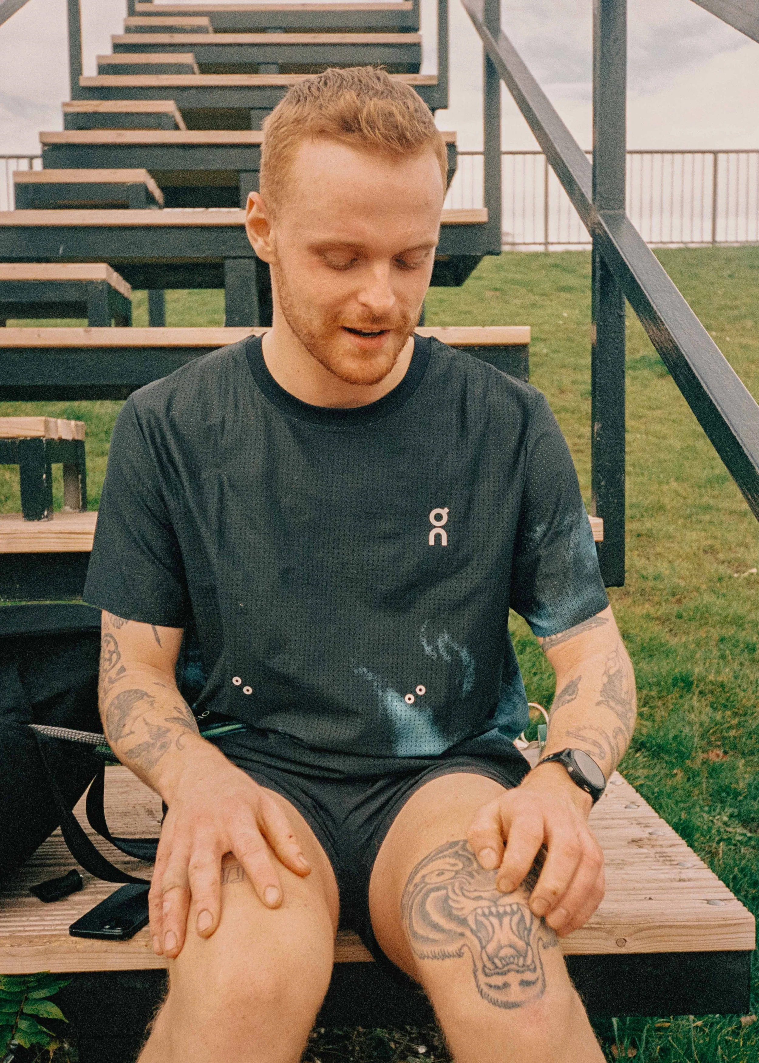 A young man sitting on a wooden bleacher, wearing a black athletic shirt with a logo, shorts, and a watch, with tattoos on his arms and leg, outdoors on a cloudy day.