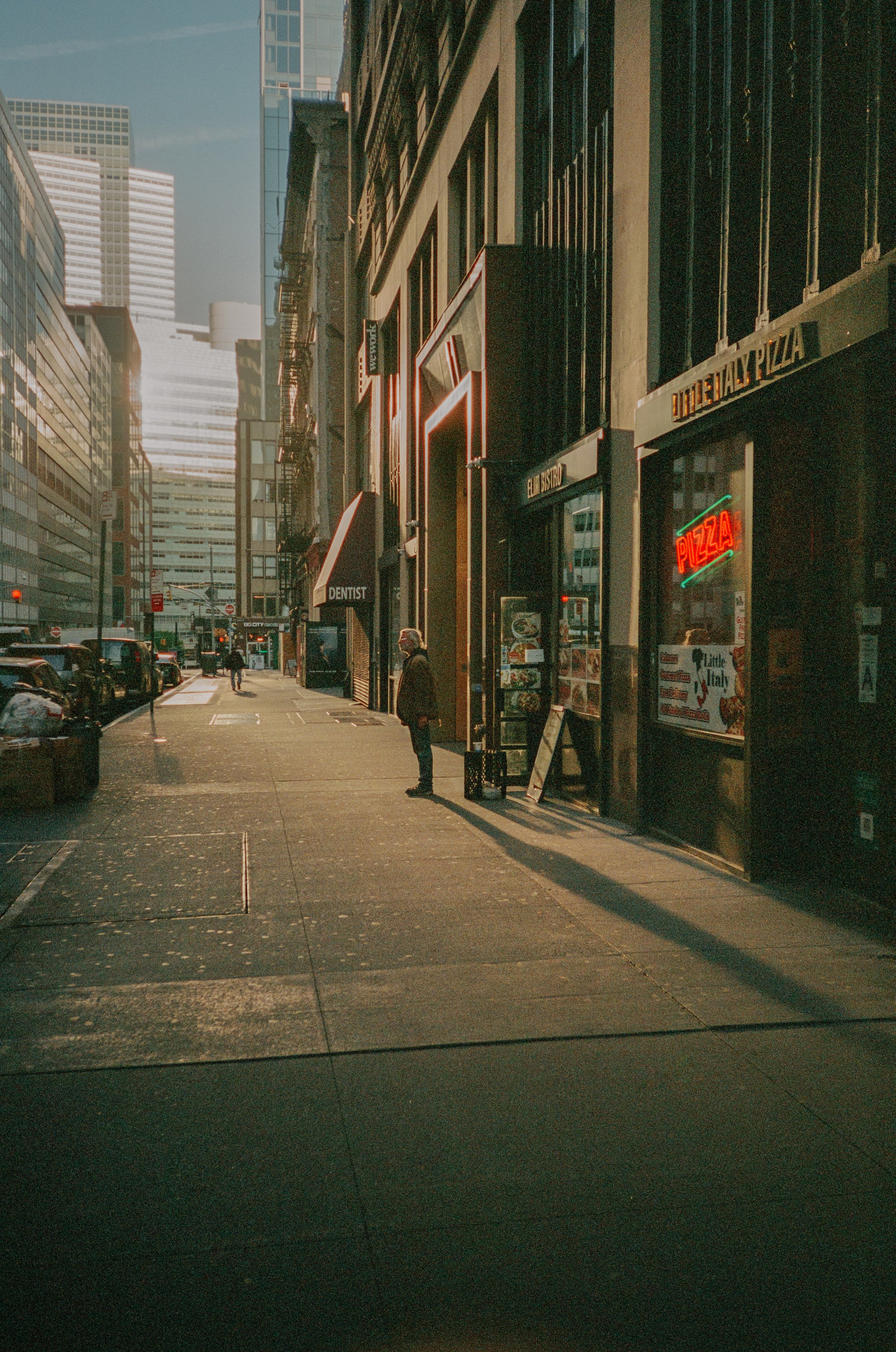 City sidewalk with people, storefronts, neon pizza sign, and tall buildings in the background on a sunny day.