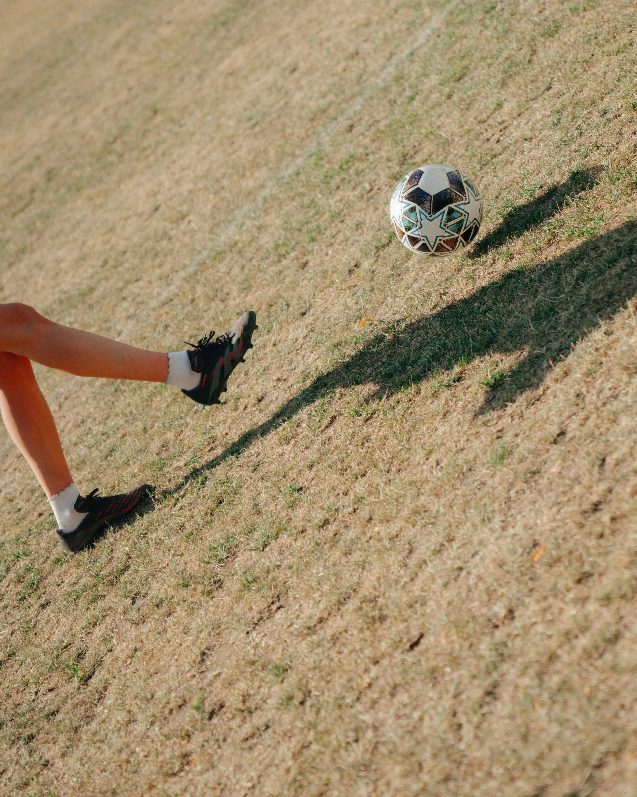 A soccer player in black cleats and white socks kicking a soccer ball on a dry, patchy grass field.