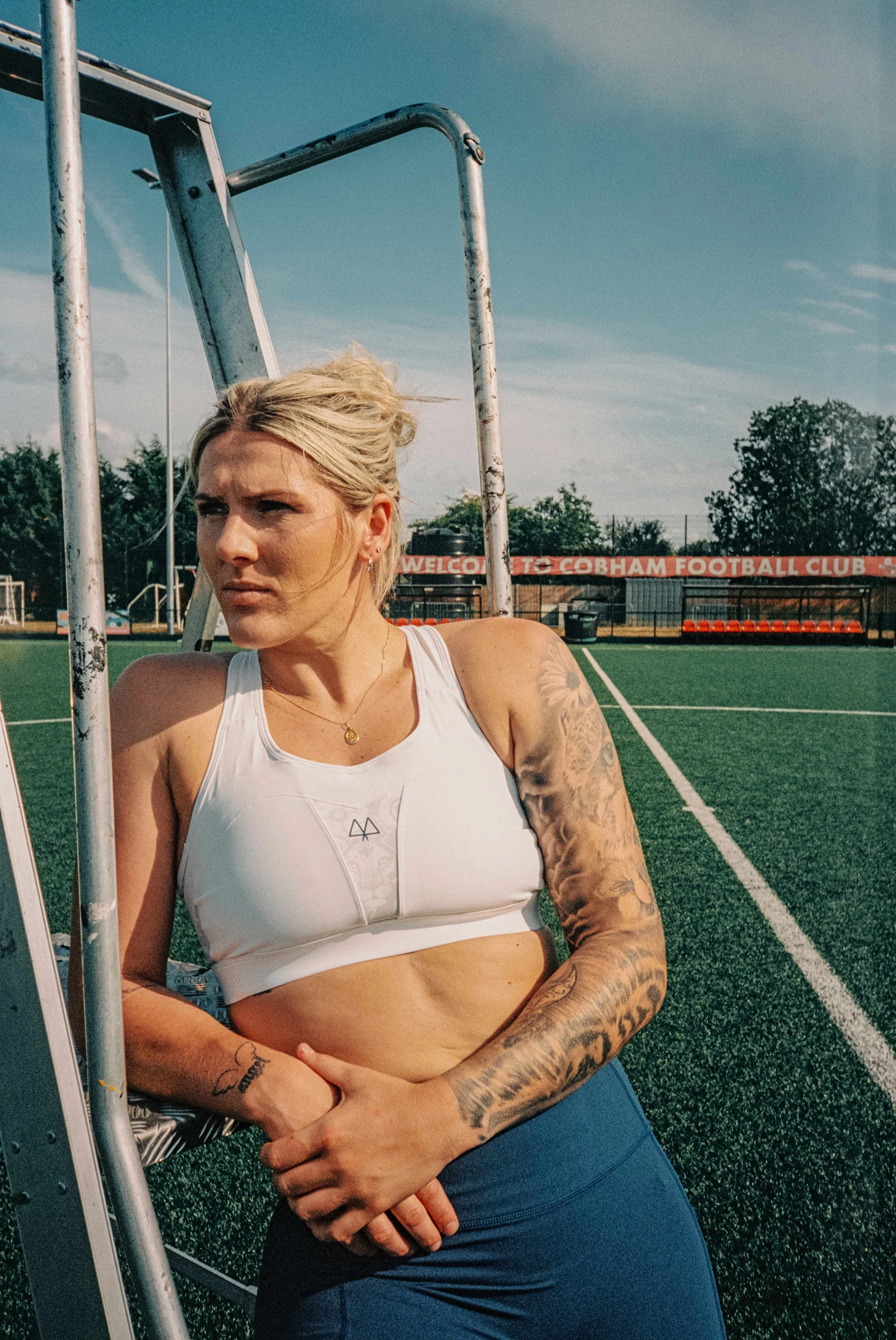 A woman with blonde hair and tattoos on her arms leaning against a soccer goal on a sports field.