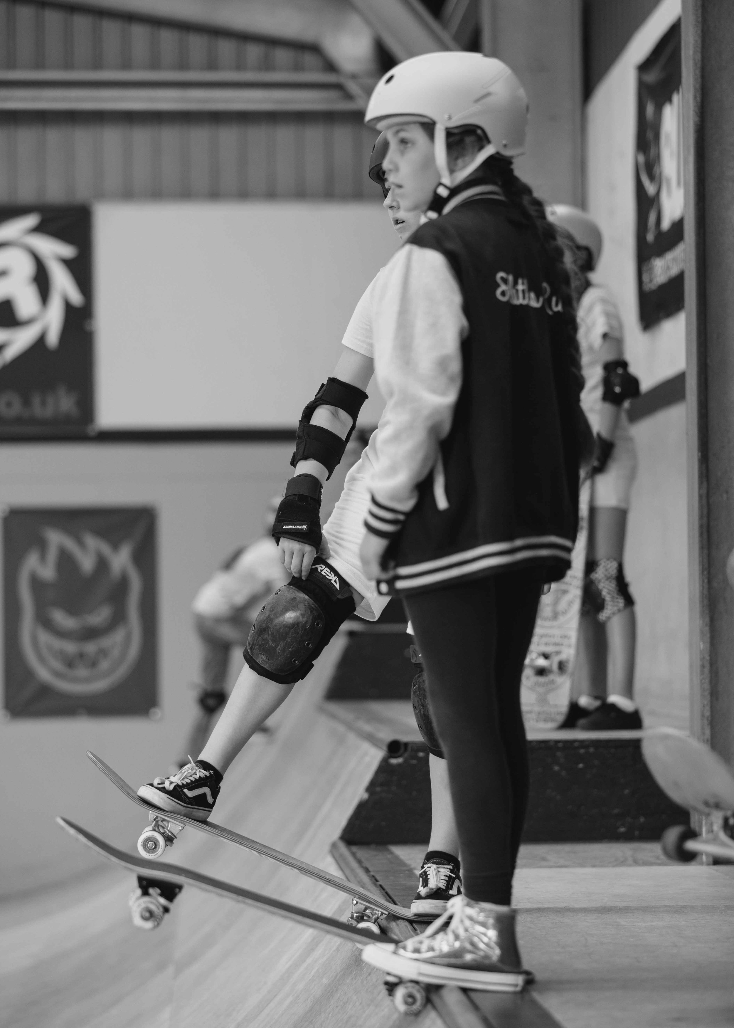 Skateboarding at an indoor skate park with a girl wearing a helmet and protective gear, standing on a ramp and holding her skateboard.