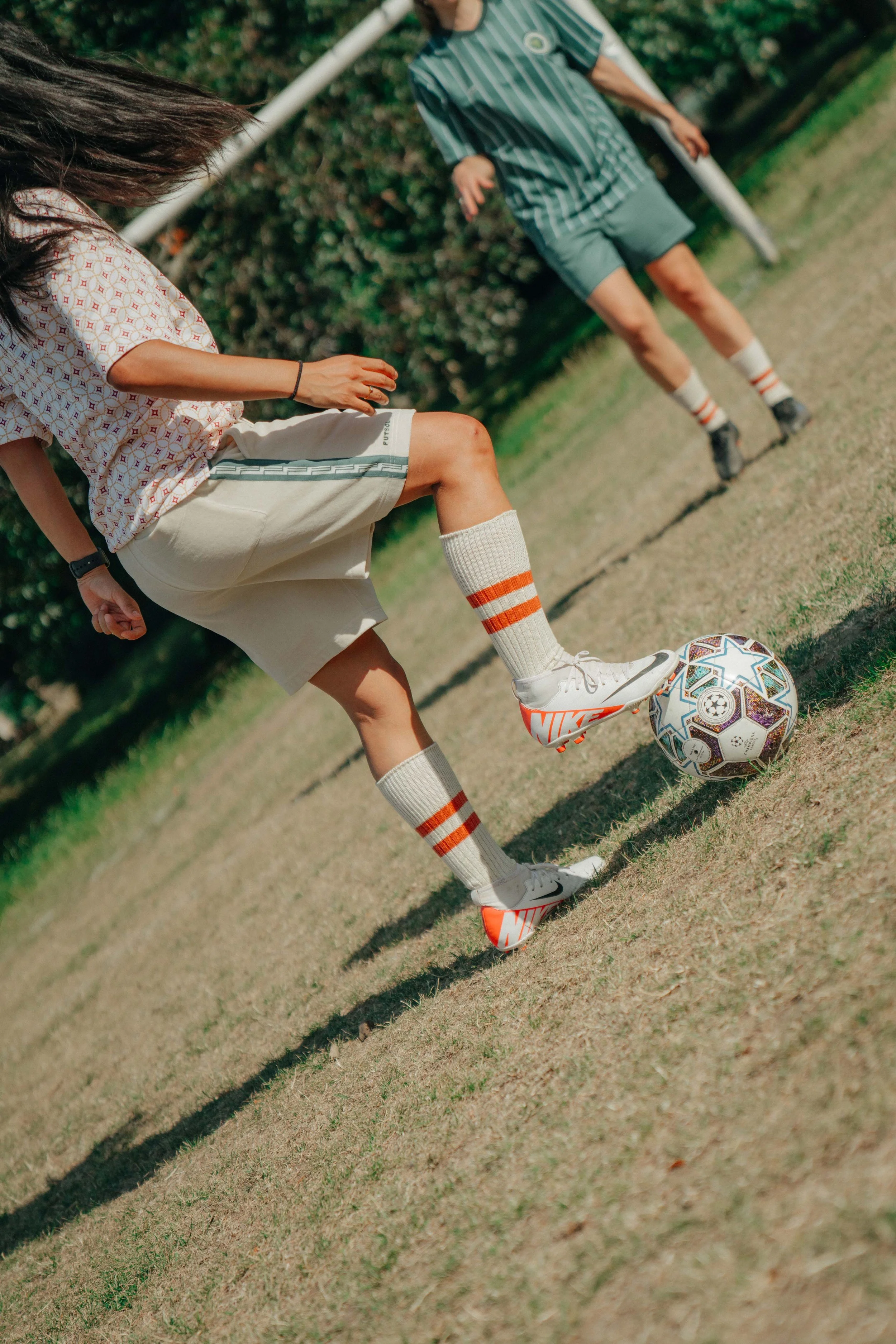 Two children playing soccer outdoors on a grassy field, with one child's foot on a soccer ball and another child in the background.