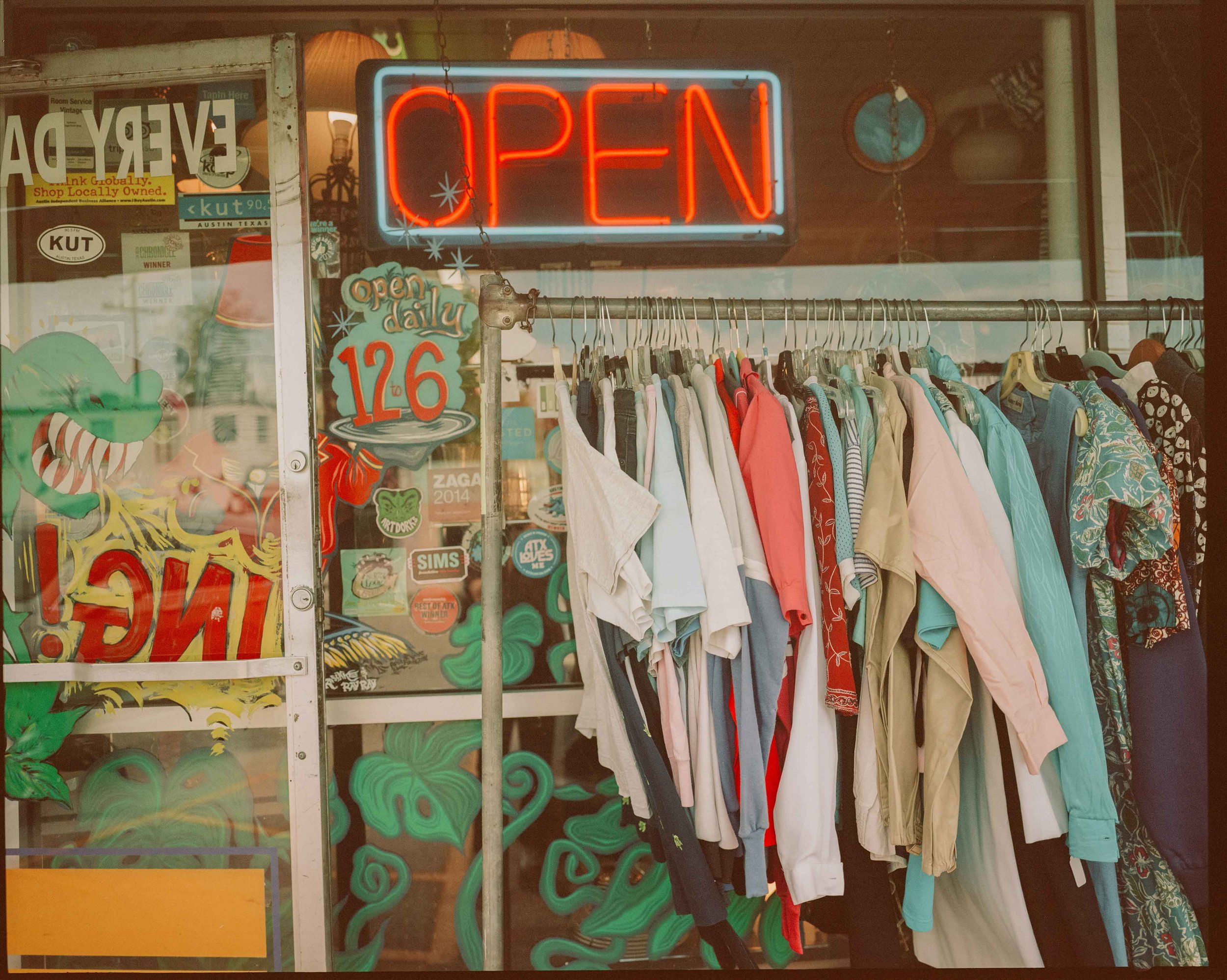 Clothing rack with various colorful shirts inside store, with an illuminated 'Open' sign and stickers on the window.