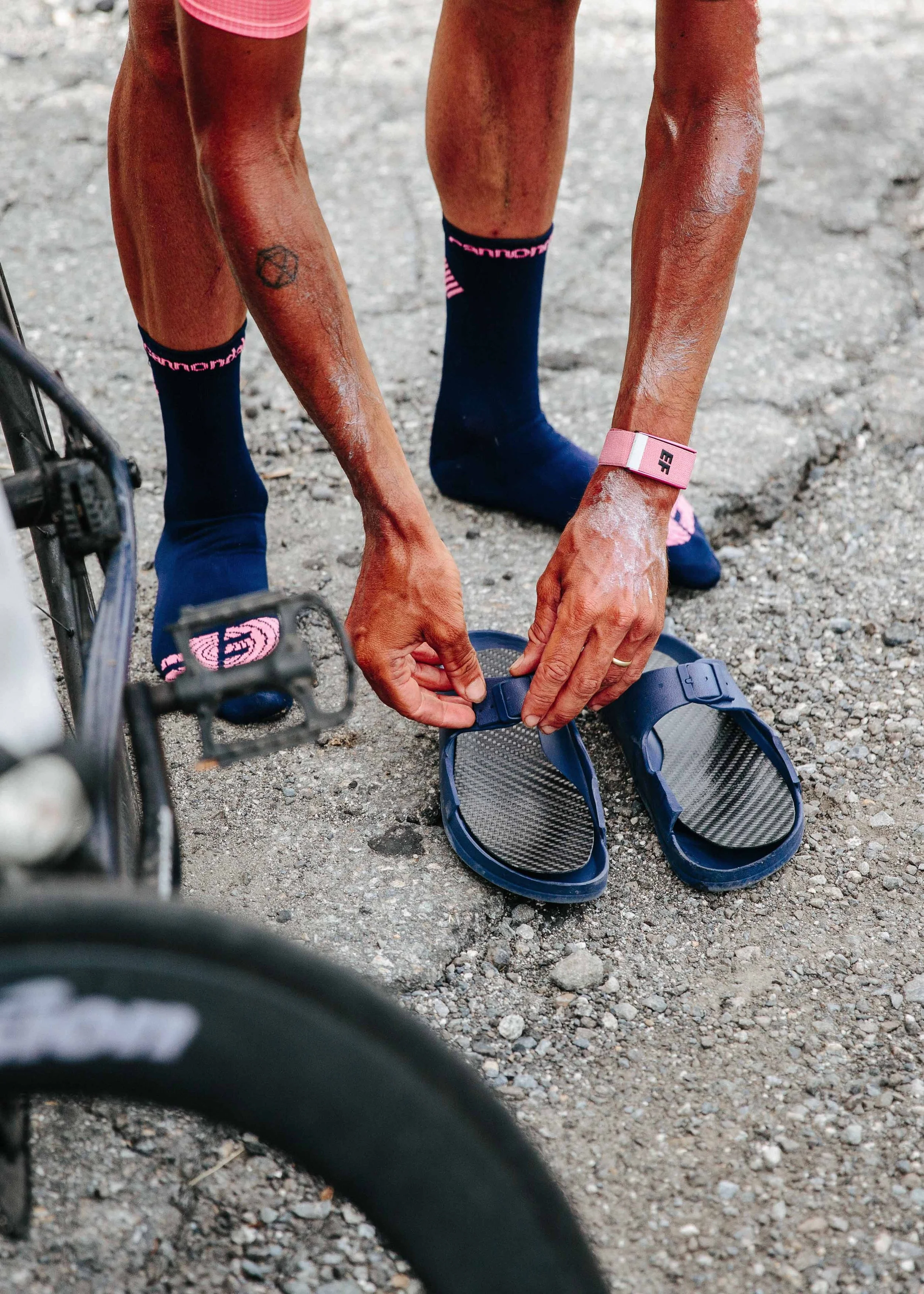 A person tying their sandal on a gravel pathway, wearing blue socks, pink shorts, and a pink fitness tracker on wrist, with a bicycle partially visible in the foreground.