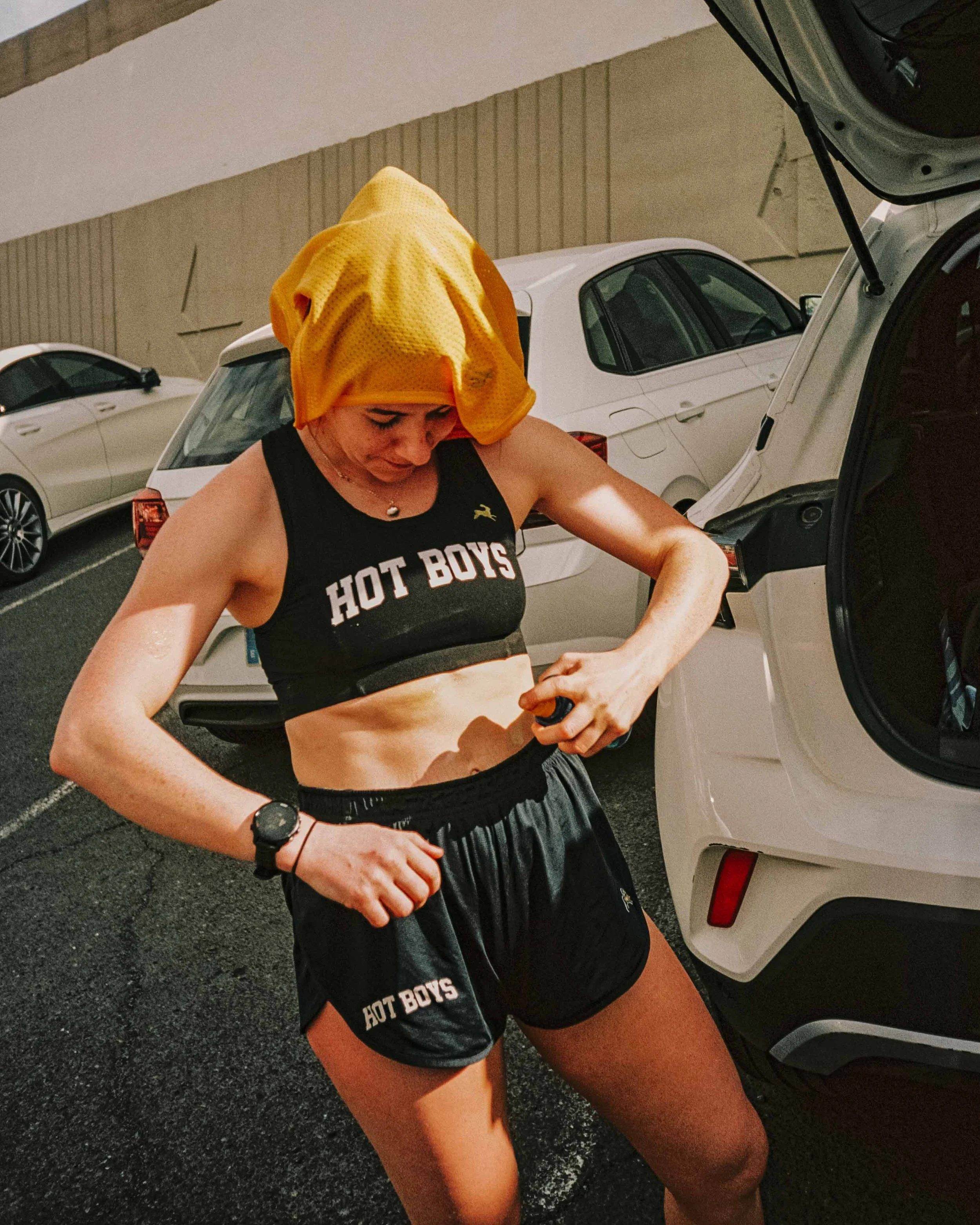 A woman in athletic clothing is standing outside near a white car, drying sweat on her arm, with her yellow towel draped over her head.