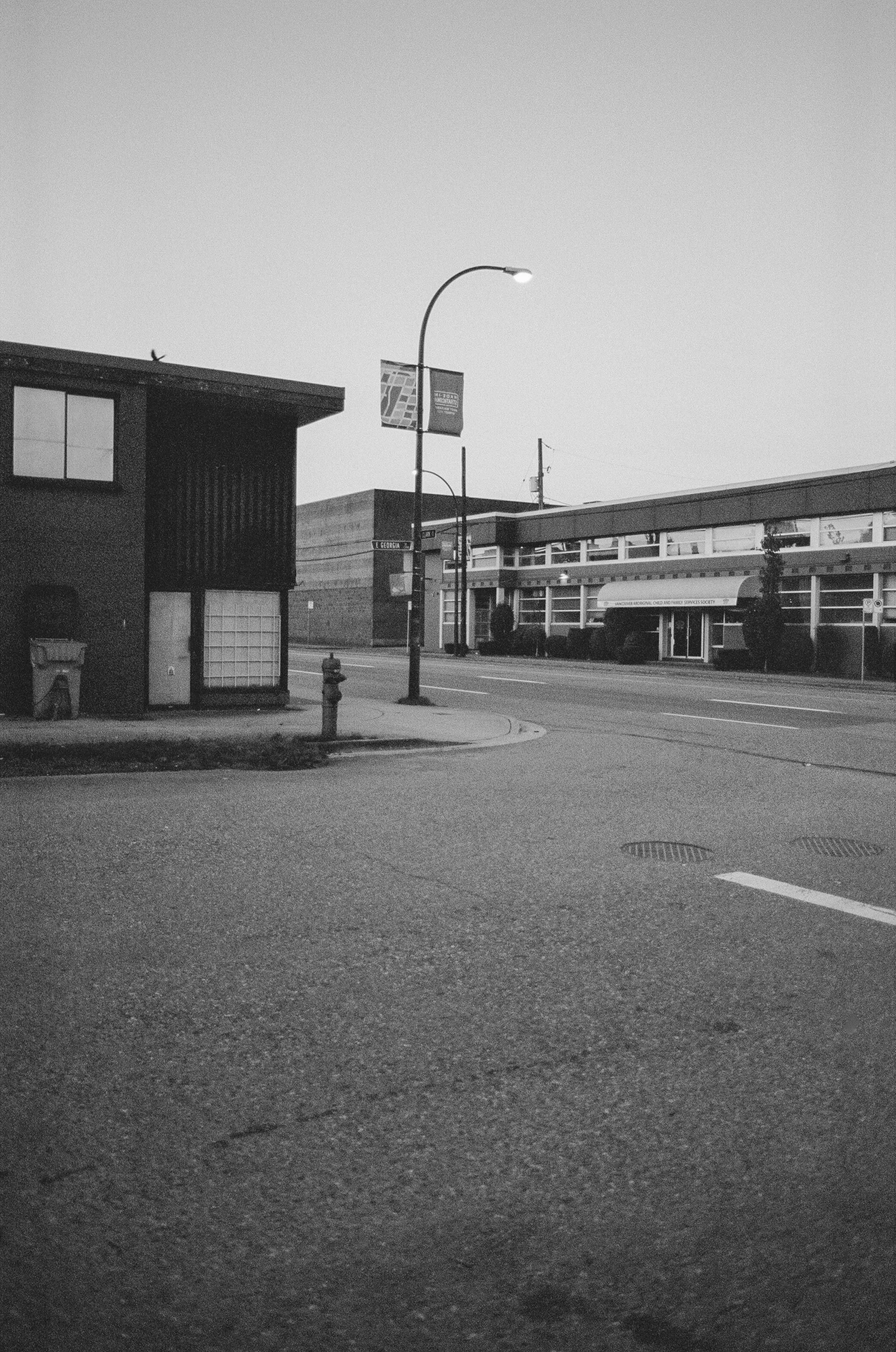 A black and white photo of a quiet urban street corner with buildings, streetlights, a sidewalk, and a fire hydrant.