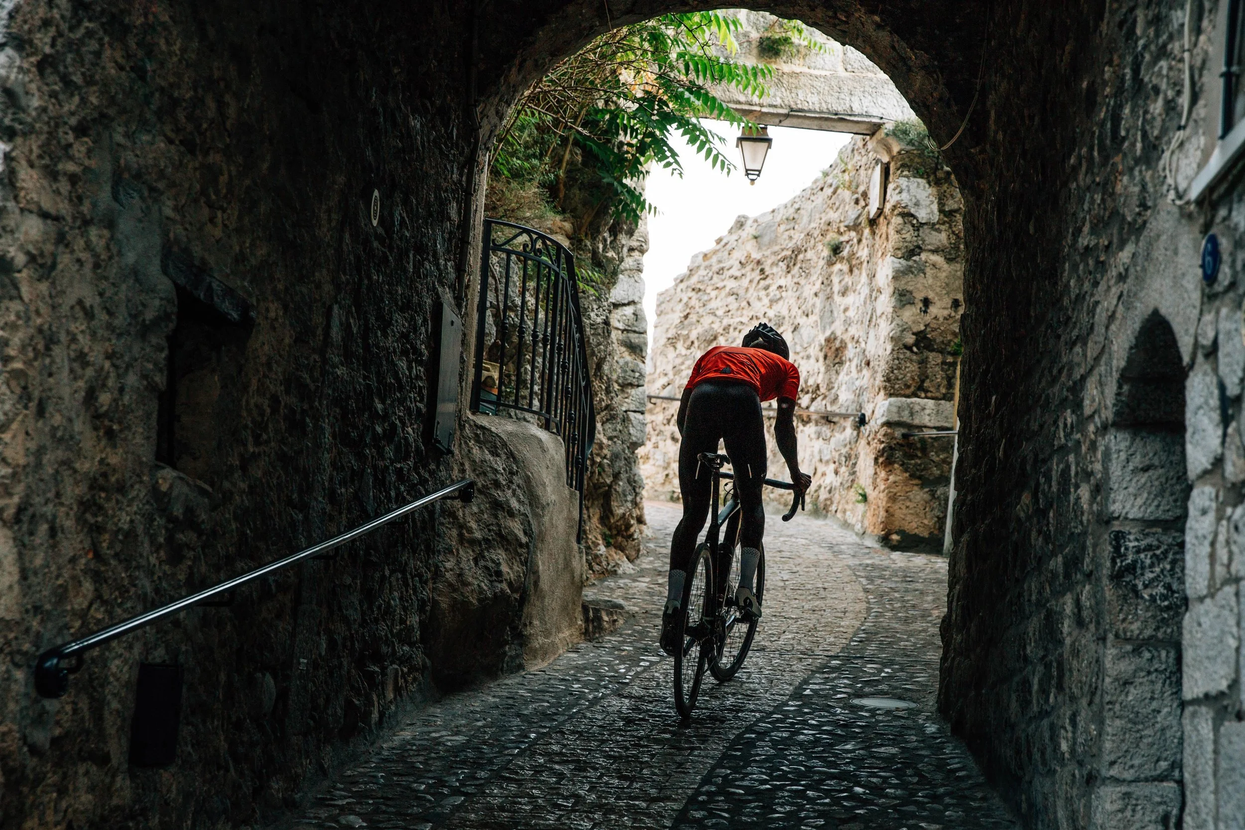 A person riding a bicycle through a narrow, cobblestone alleyway with stone walls and a stone arch overhead, illuminated by a street lamp.