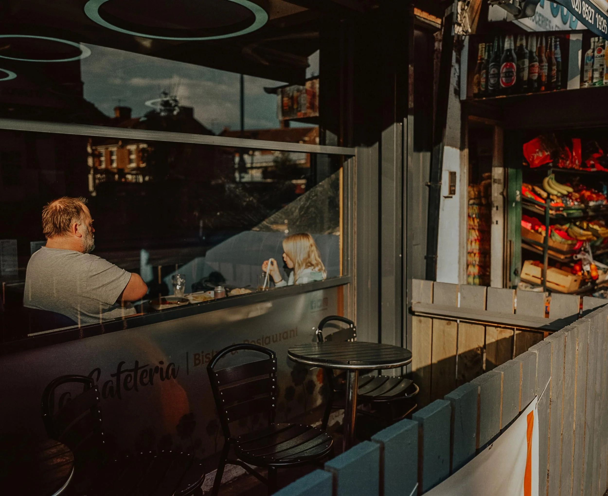 View through a restaurant window showing a man and a woman eating inside, with outdoor chairs and tables in front, and a store with snacks and bananas visible to the right.