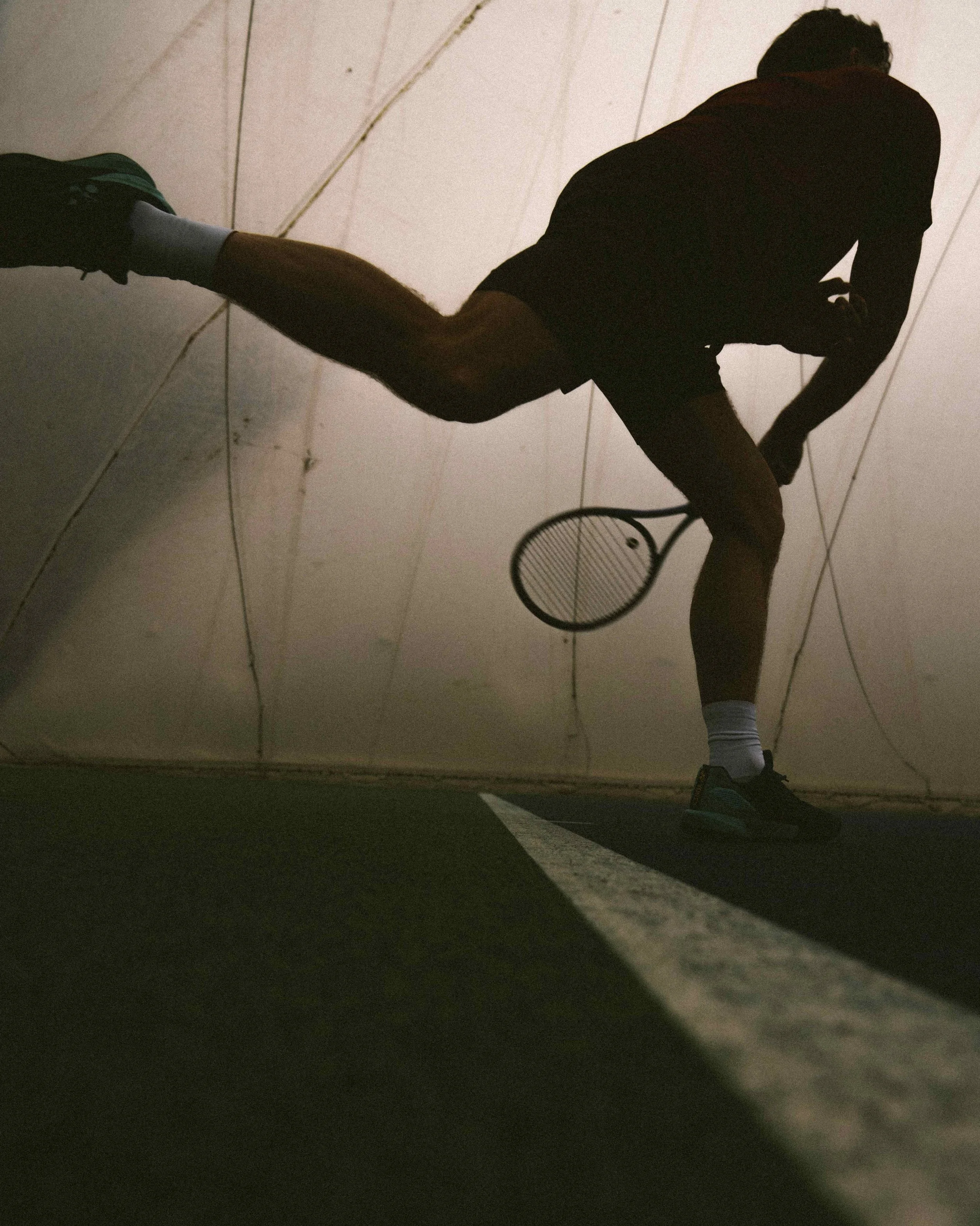 Silhouette of a male tennis player in a court, holding a tennis racket, preparing to hit a shot, with a low camera angle capturing the scene.