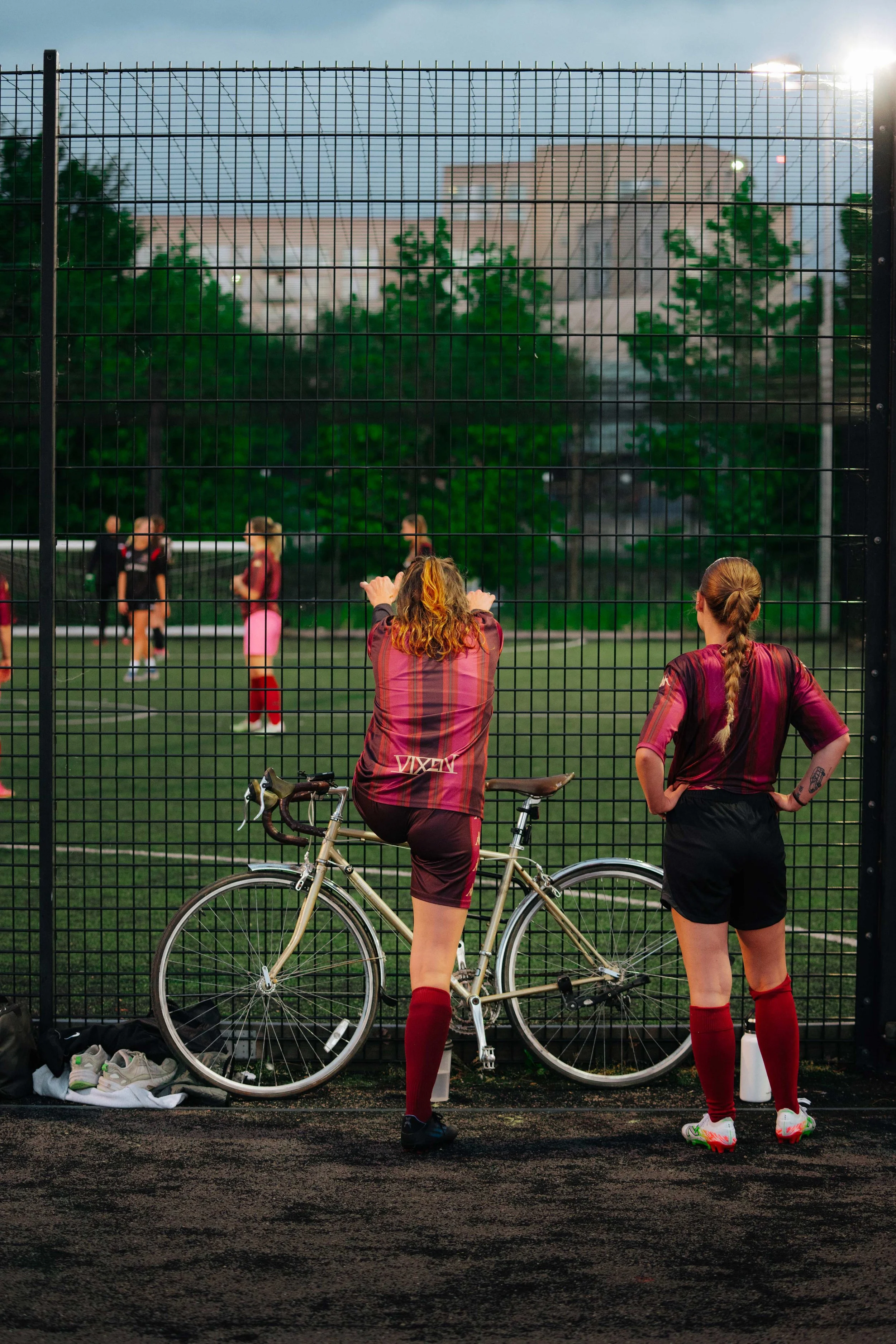 Two female soccer players in maroon and black uniforms standing near a bicycle, watching a game on a fenced soccer field with multiple players, trees, and buildings in the background.