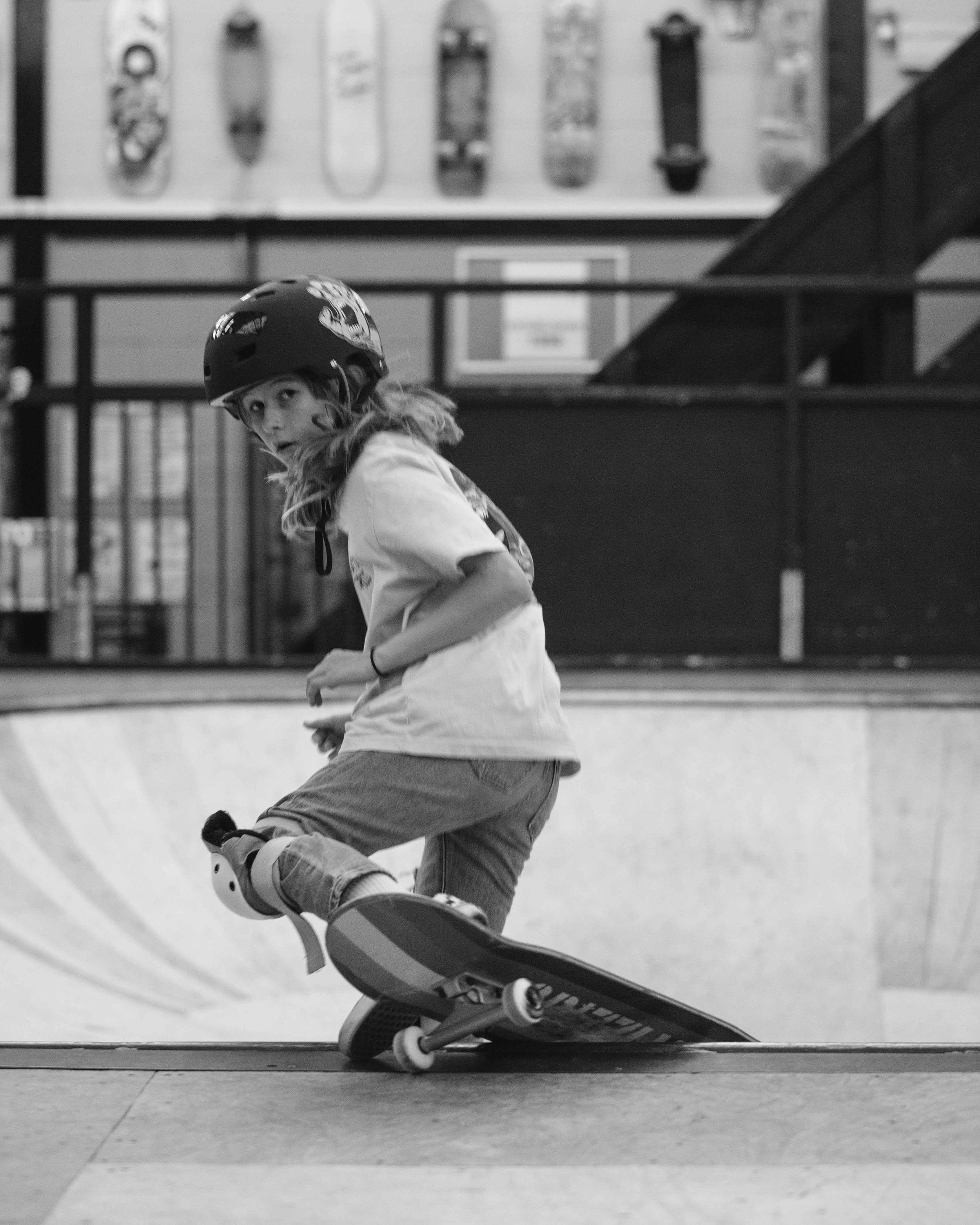 A young girl skateboarding at an indoor skate park, wearing a helmet and casual clothes, with skateboards displayed on the wall behind her.