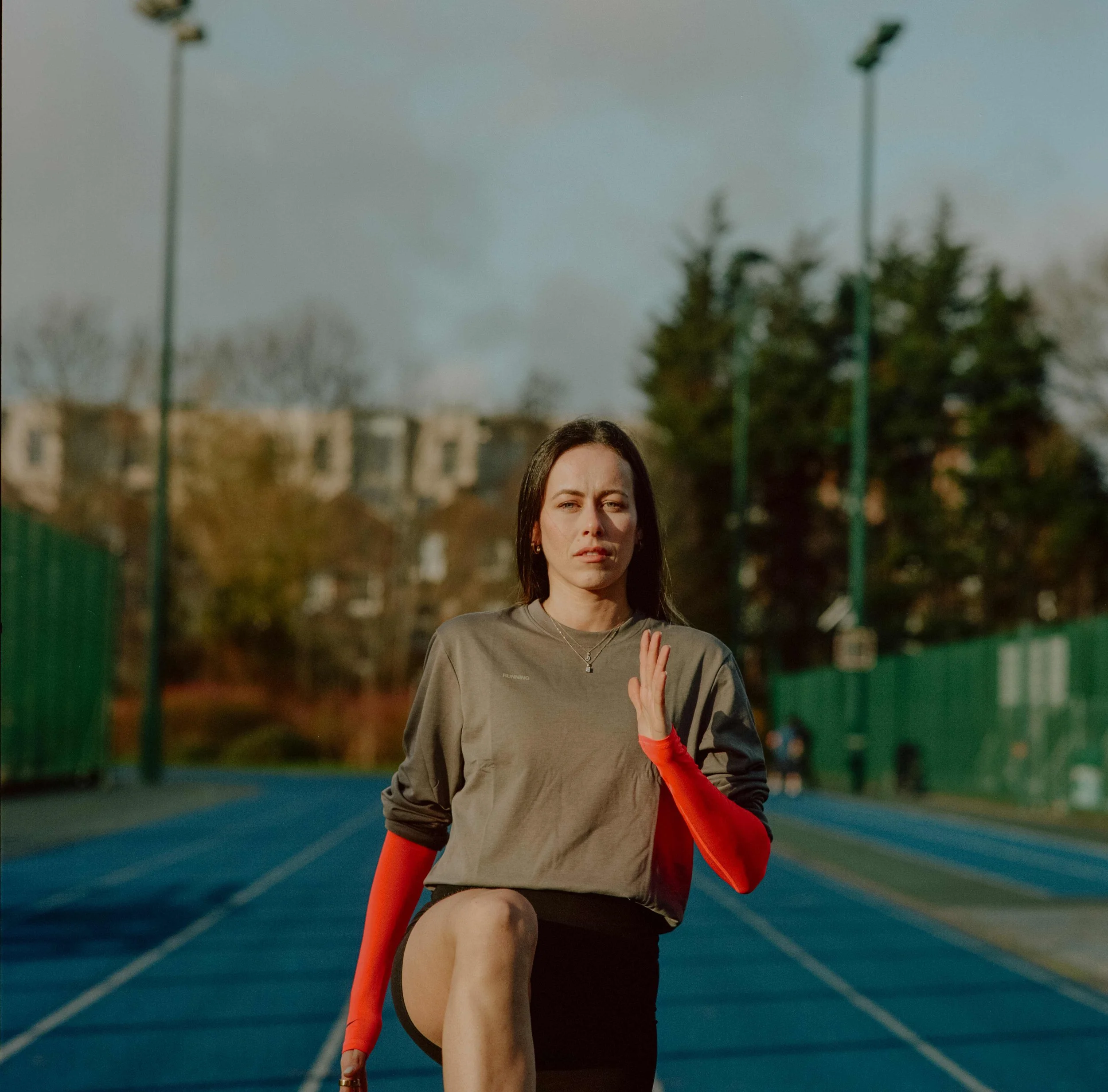 A woman running on a track, wearing a gray sweatshirt with orange sleeves and black shorts, with focused expression, set against an outdoor athletic field and cloudy sky.