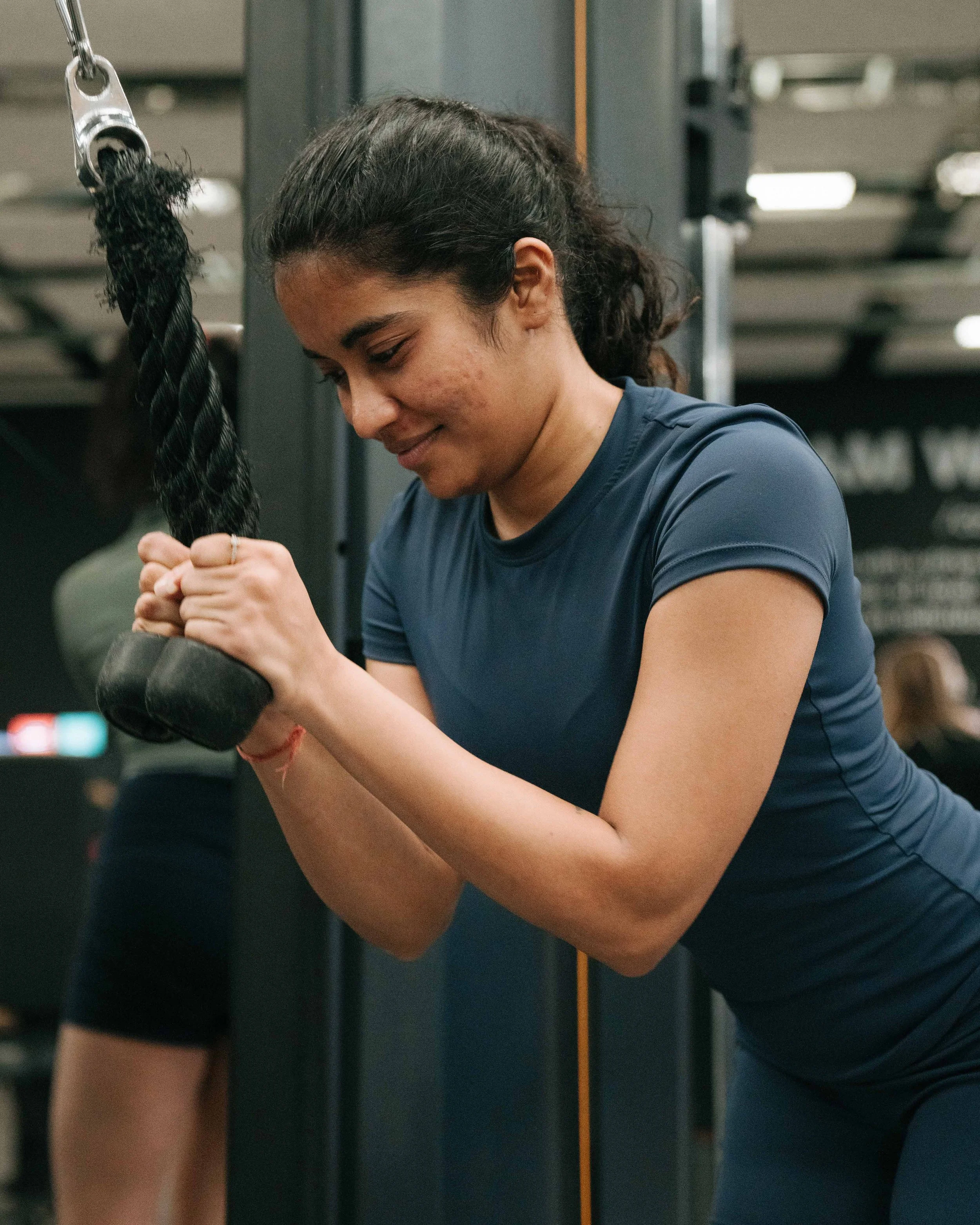 A woman in a blue athletic shirt is doing a tricep pushdown exercise in a gym.