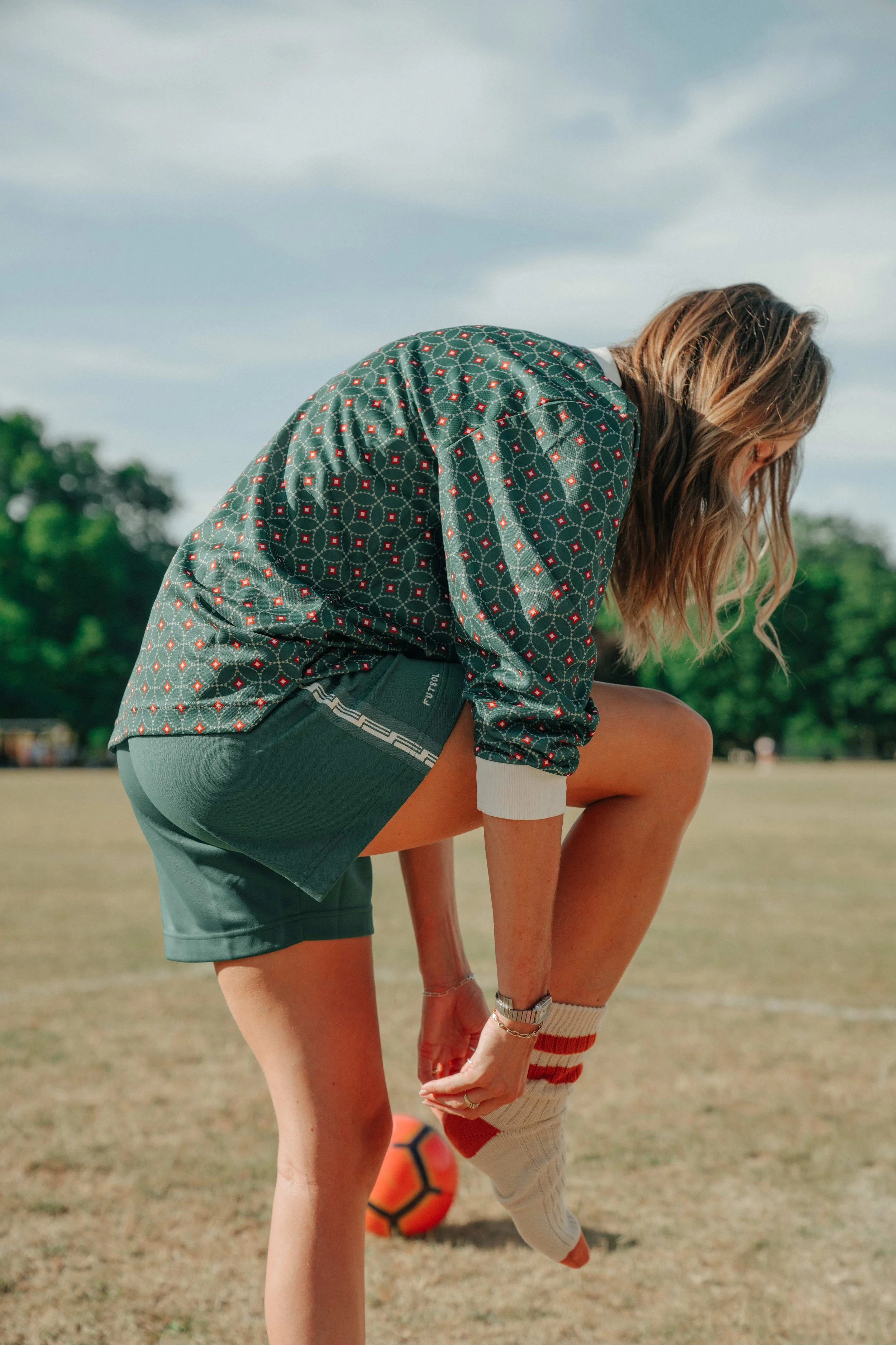 A woman outdoors on a grassy field, stretching or adjusting her sock before playing soccer, with a soccer ball in the background and trees under a partly cloudy sky.