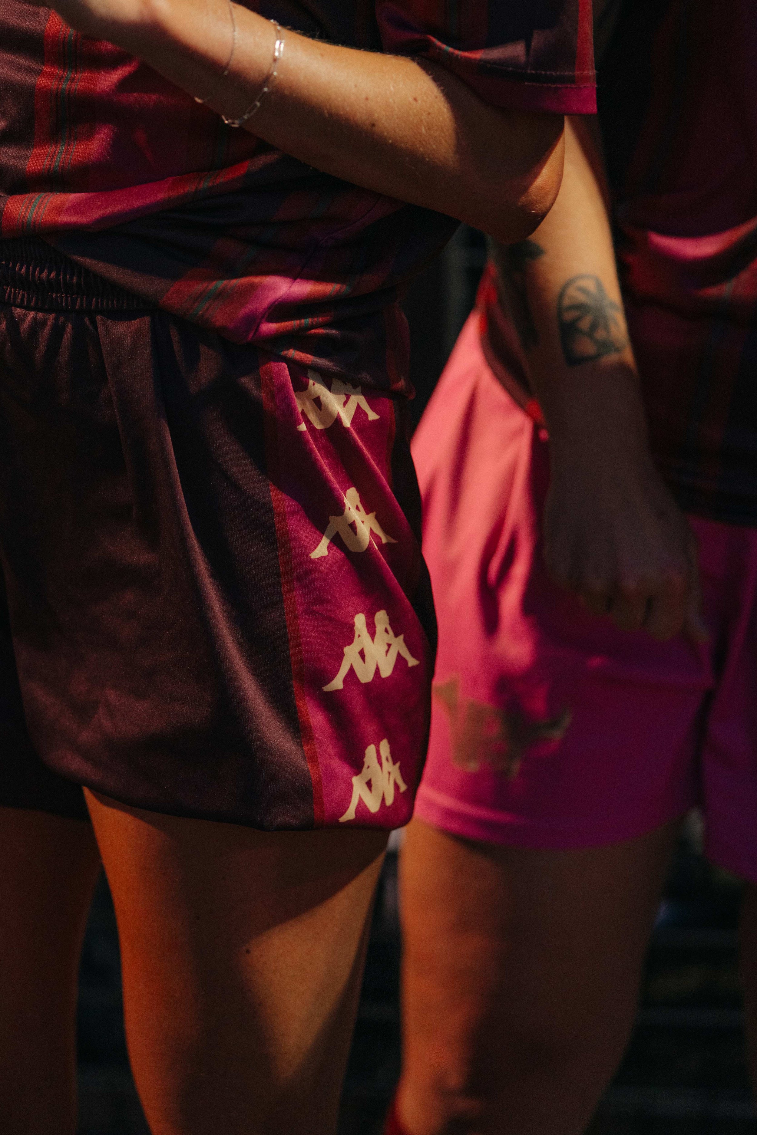Close-up of two people wearing sports shorts, one with black and maroon Kappa shorts and the other with pink shorts, with tattoos visible on their arms.
