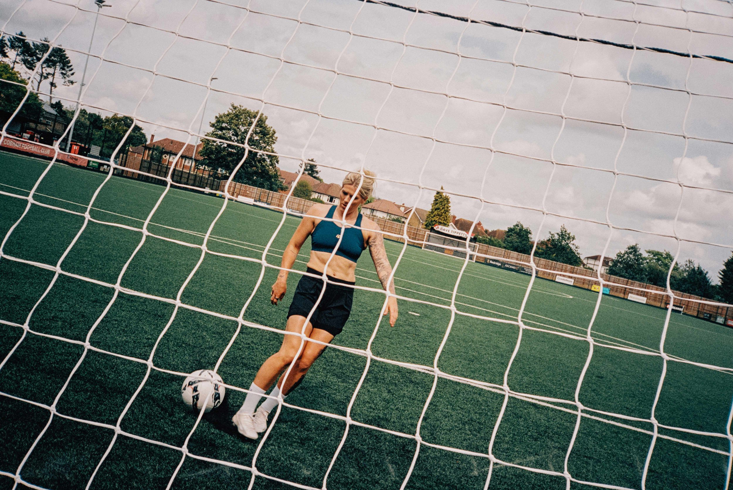 A woman with blonde hair, wearing a blue sports bra and black shorts, is standing on a soccer field with a soccer ball near her foot, viewed through a goal net.