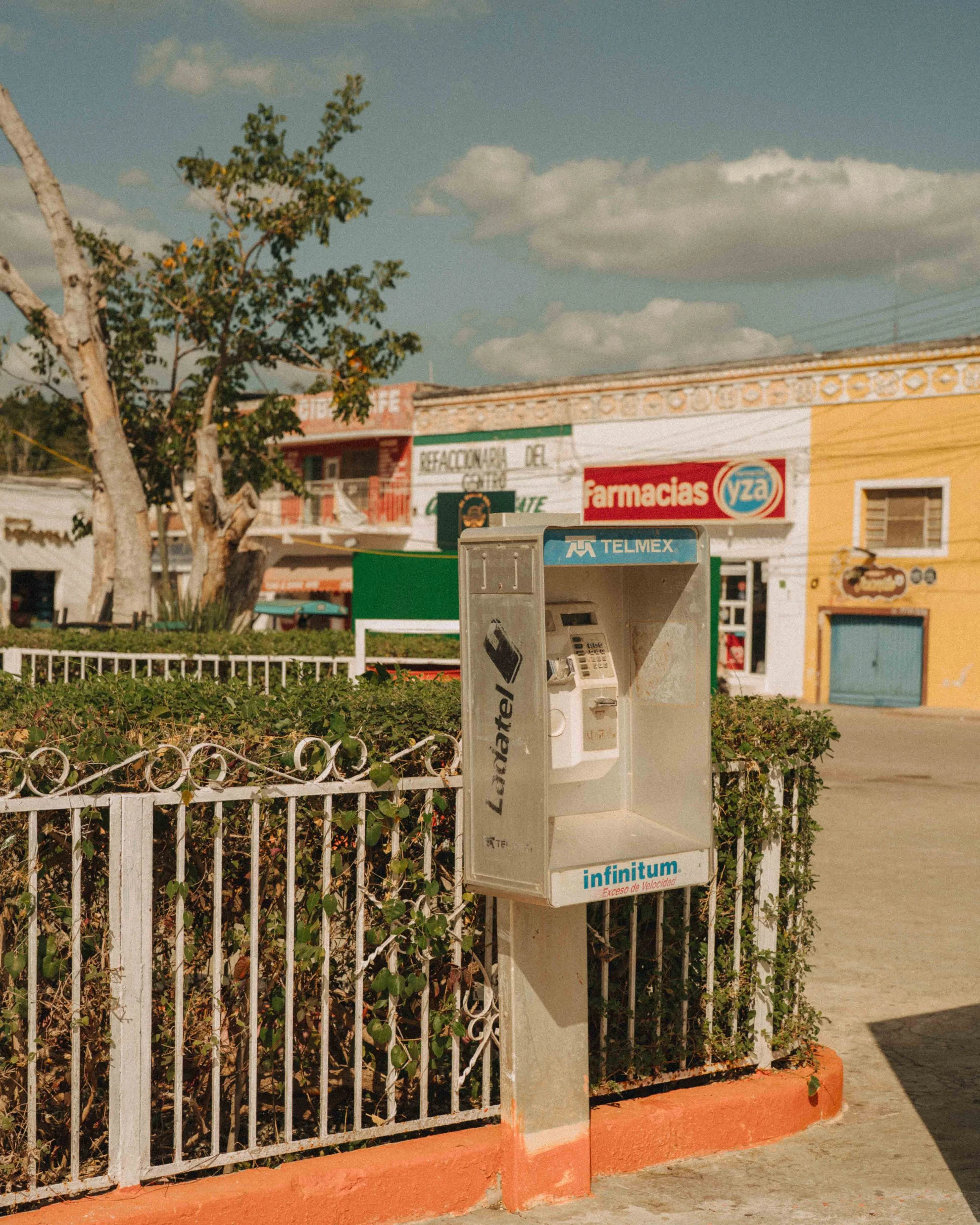 Payphone on a sidewalk in a small town, with shops and colorful buildings in the background, under a partly cloudy sky.