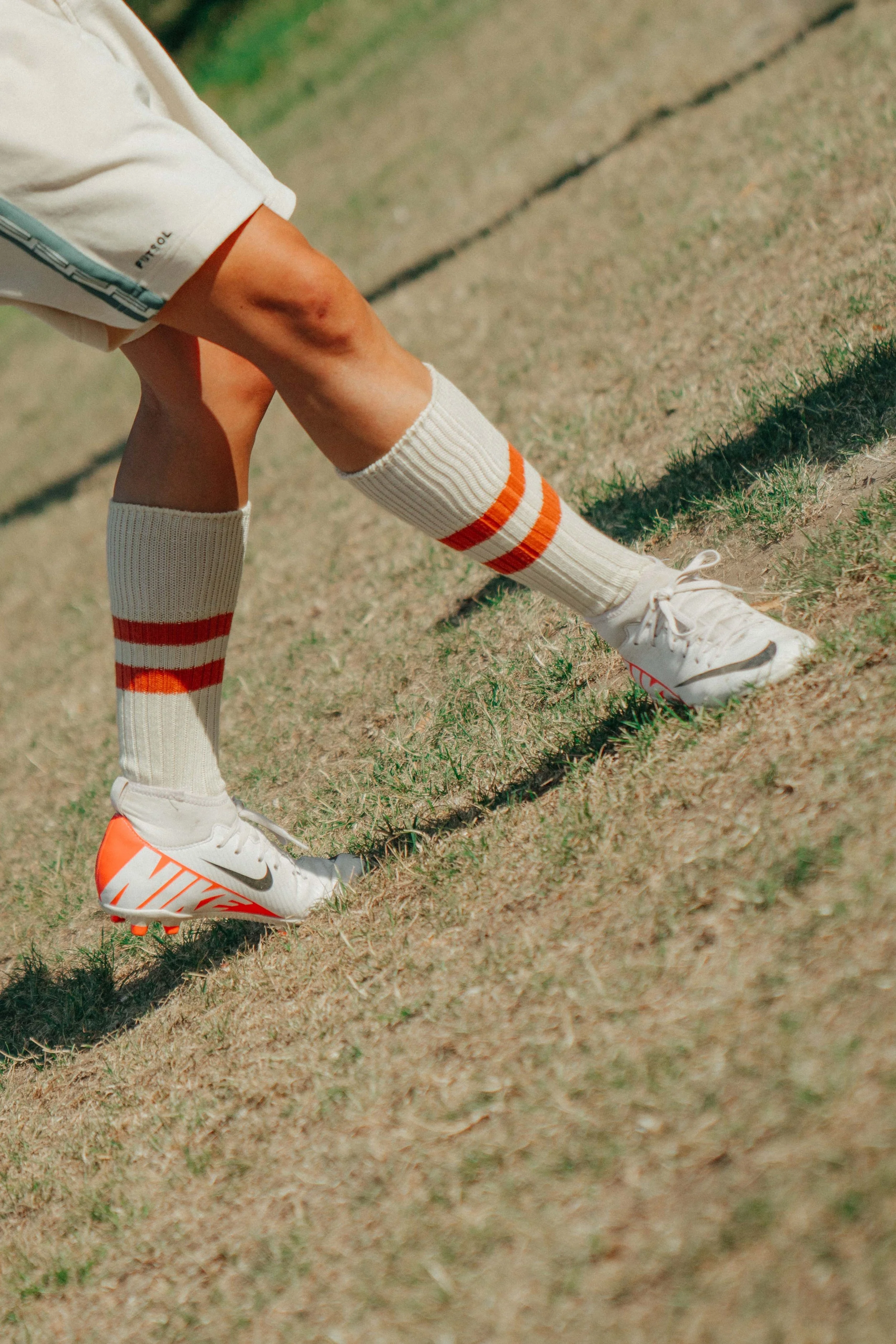 A person wearing white Nike cleats, white knee-high socks with orange stripes, and beige shorts is in a crouched position on a grassy field, likely preparing for a sports activity.