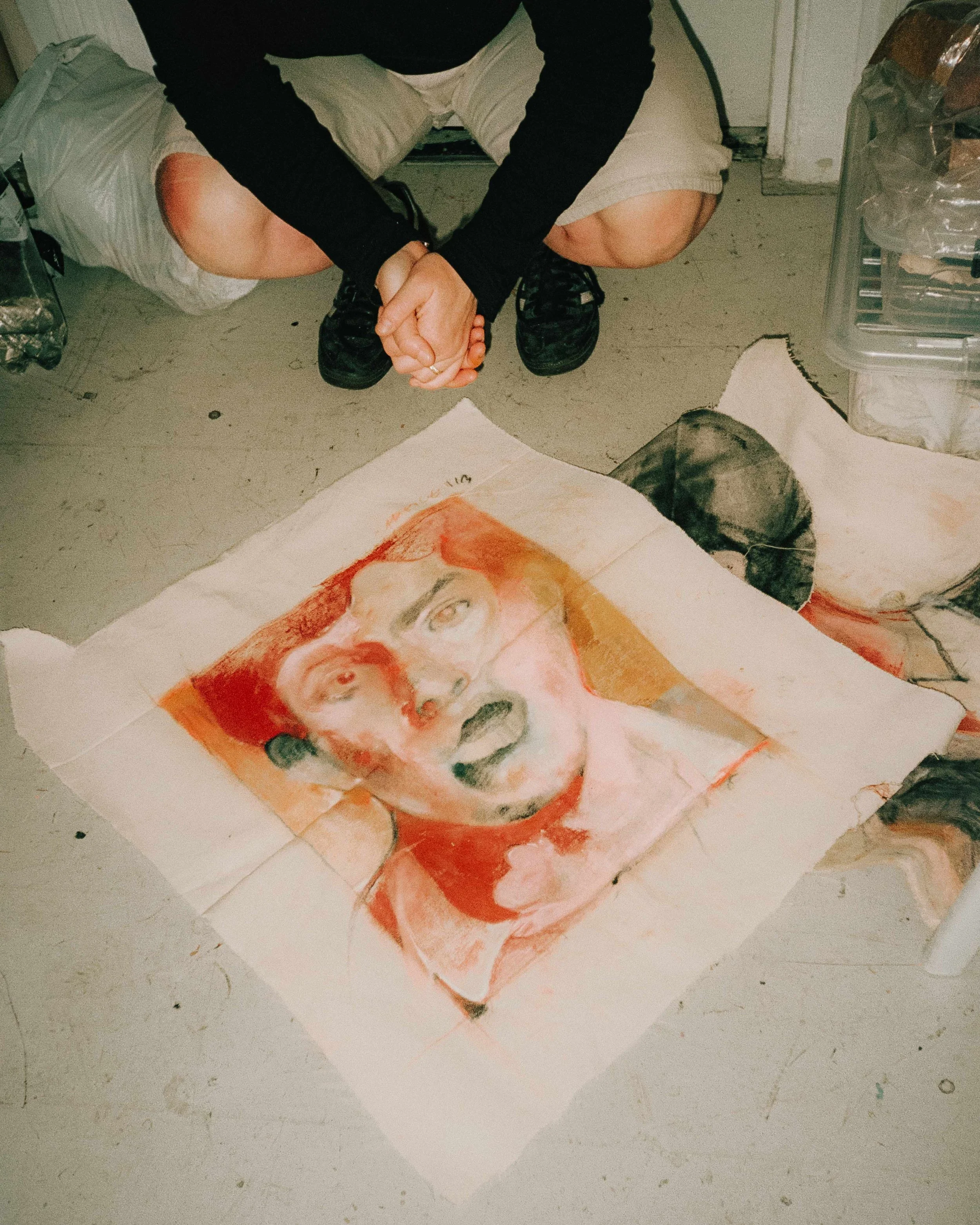 A person with their head and shoulders visible kneeling on the floor, wearing beige shorts and a black top, with hands clasped. In front of them is a large watercolor or pastel painting of a person's face on a piece of paper, depicting an expressive, surprised or shocked expression with open mouth and wide eyes.