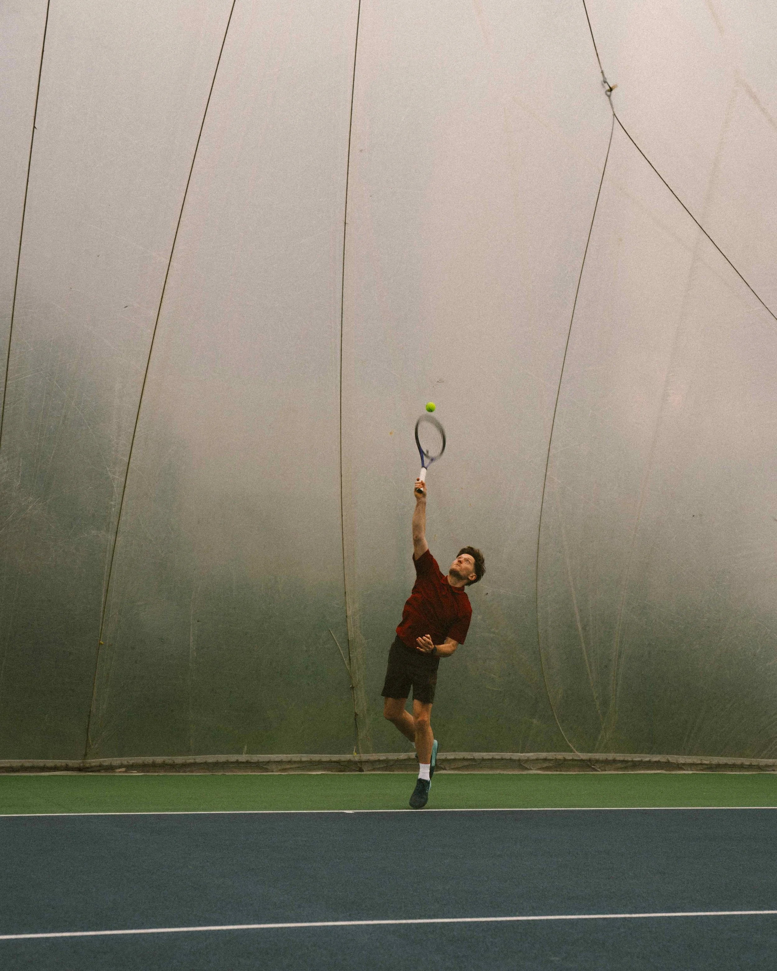 A man playing tennis indoors, reaching to hit a tennis ball with his racket, on a court with green and black flooring, with a gray wall in the background.