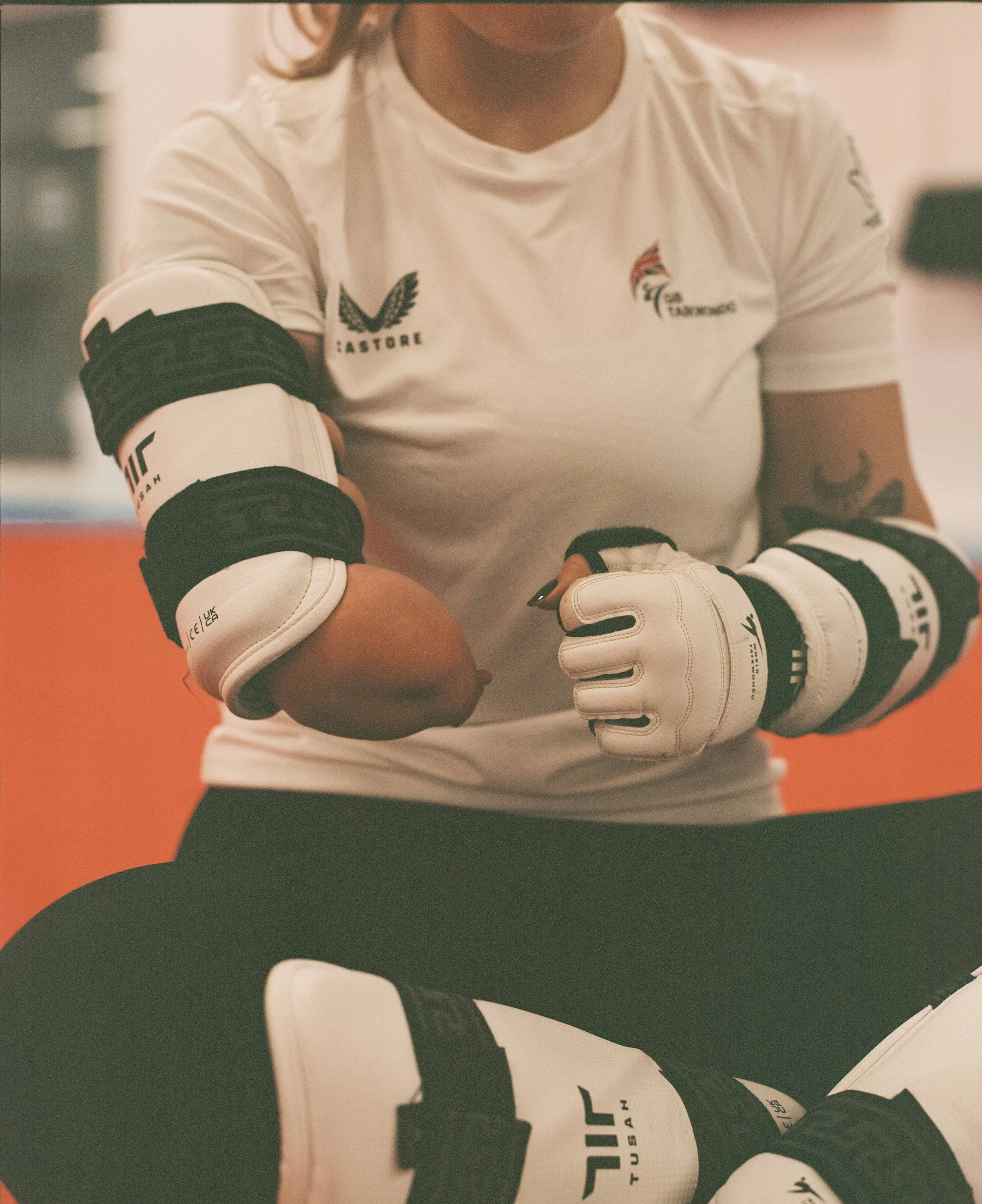 A mixed martial arts fighter prepares for a match in a gym, wearing white fight gloves, with logos on her shirt and gear.