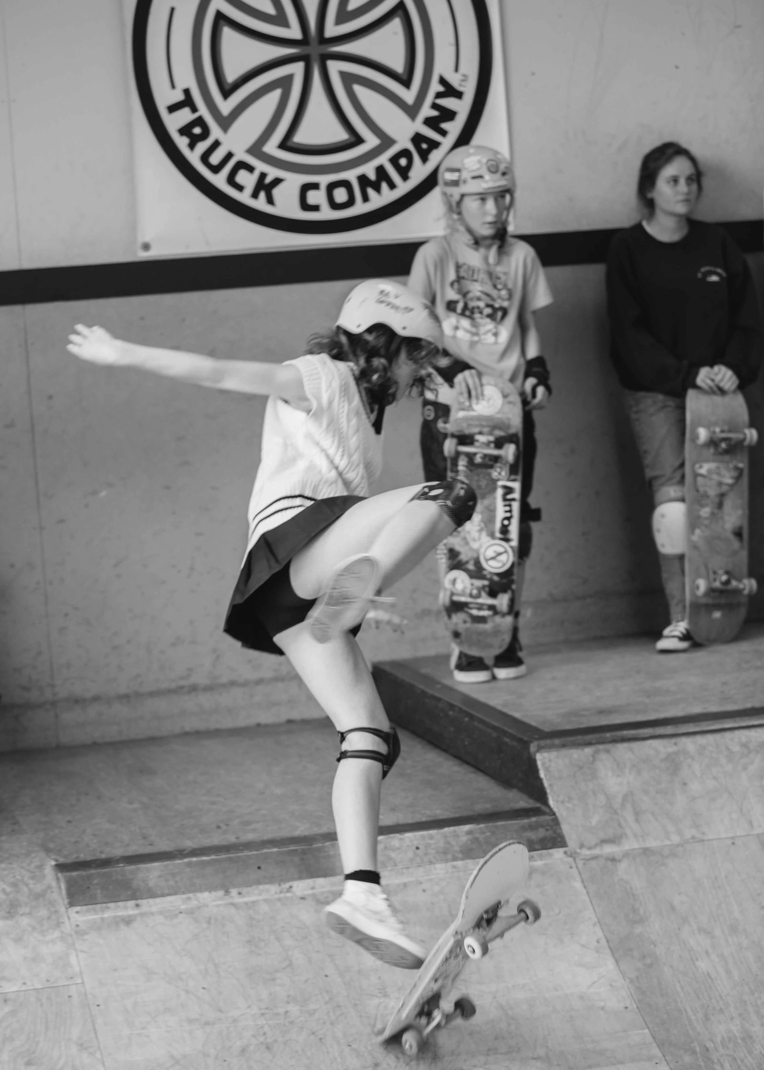 A young girl is skateboarding at an indoor skate park, wearing a helmet and knee pads, with two other girls holding skateboards in the background.