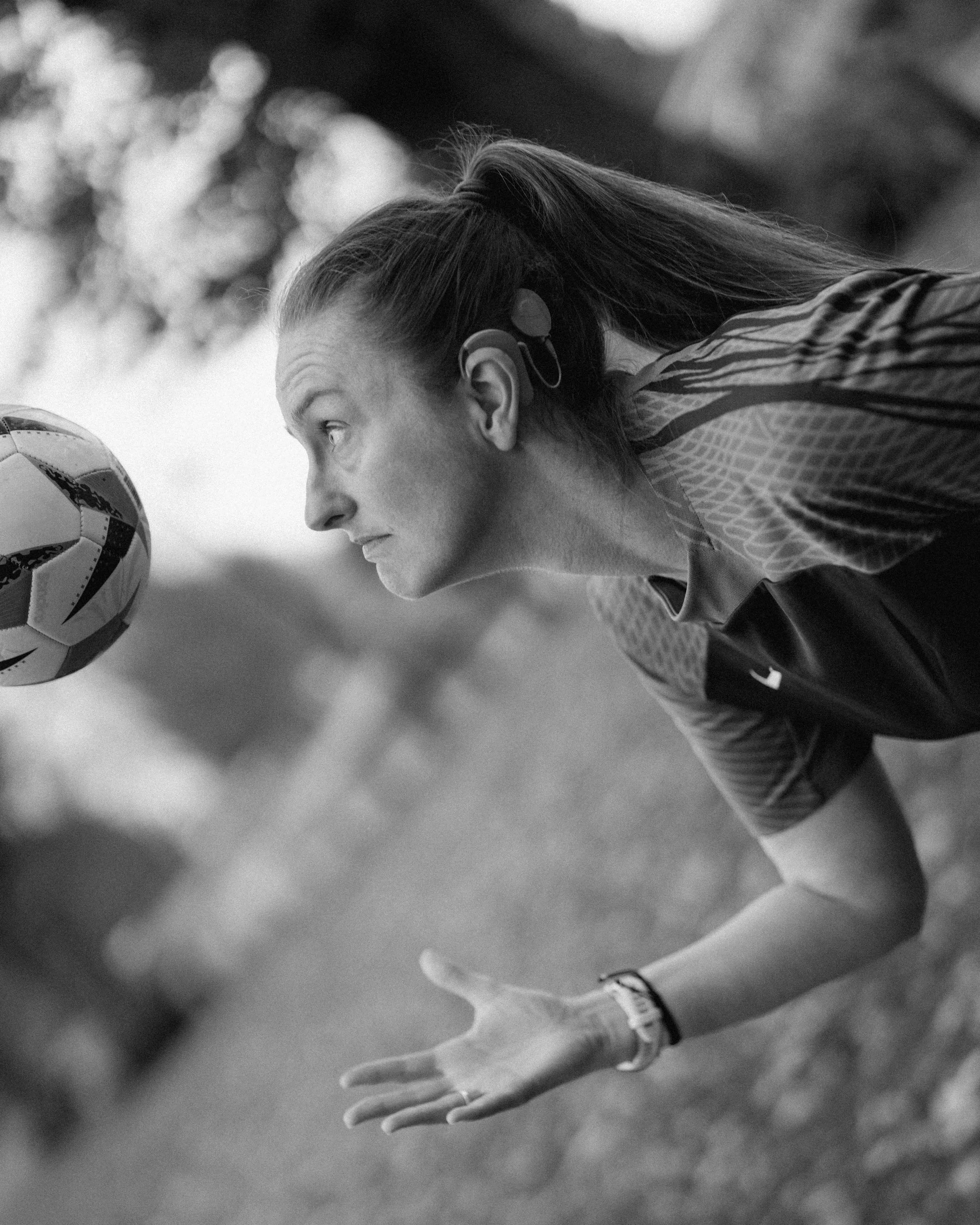A woman outdoors, wearing sports gear and a hearing aid, preparing to head a soccer ball during a game.