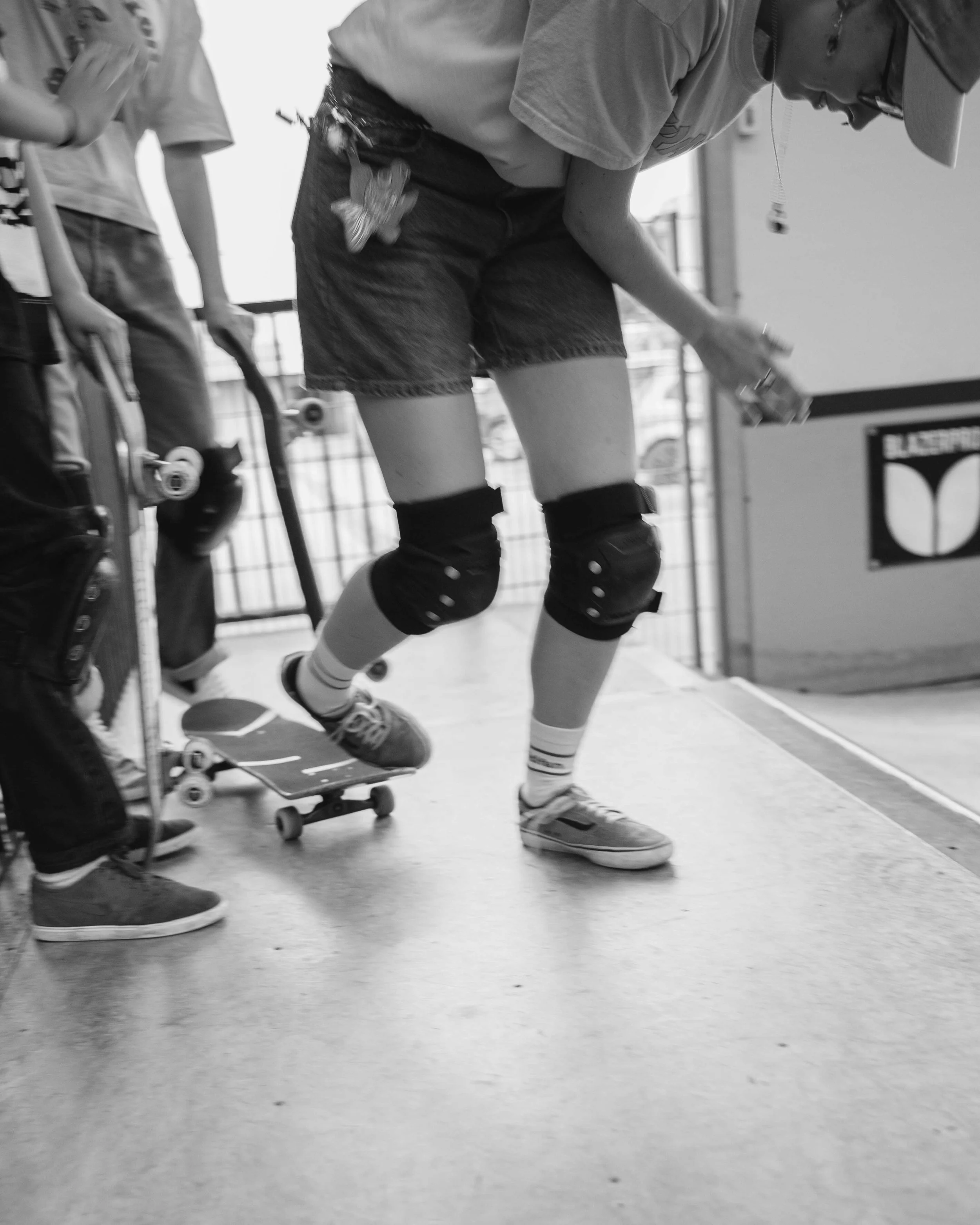 A person wearing knee pads skateboarding indoors, with another person standing nearby holding a skateboard.