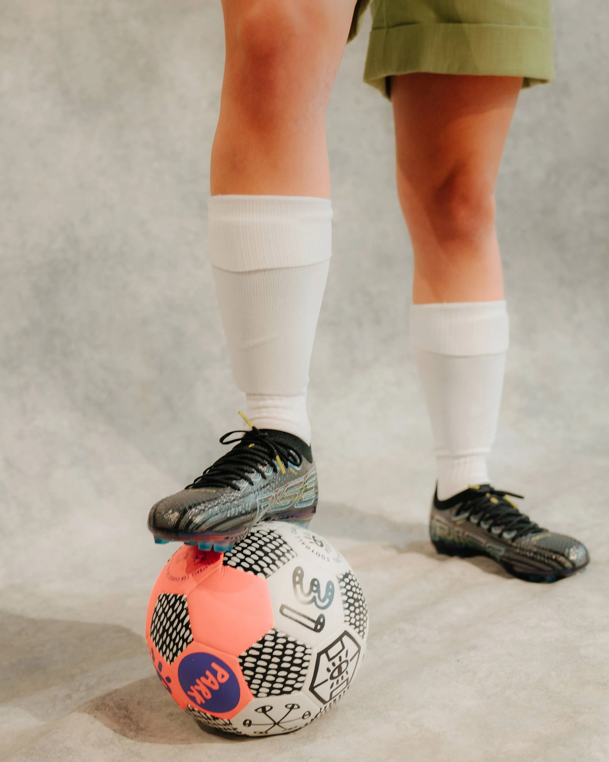 A person in athletic shoes and white socks stands with one foot on a colorful soccer ball inside a neutral-colored room.