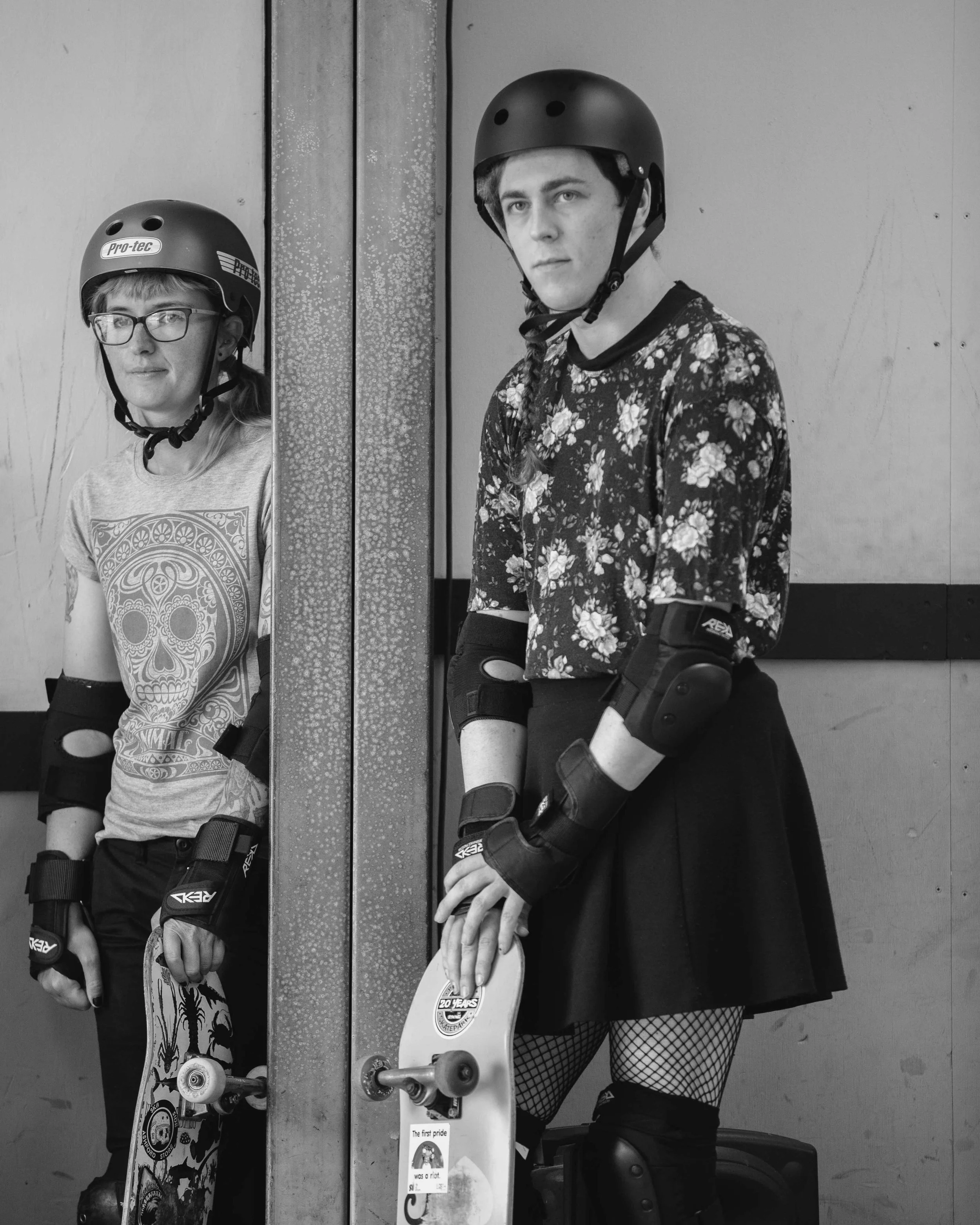 Two young women wearing skate helmets and protective gear, standing next to a vertical metal post. One is holding a skateboard, the other holding a helmet, both looking at the camera. The image is black and white.