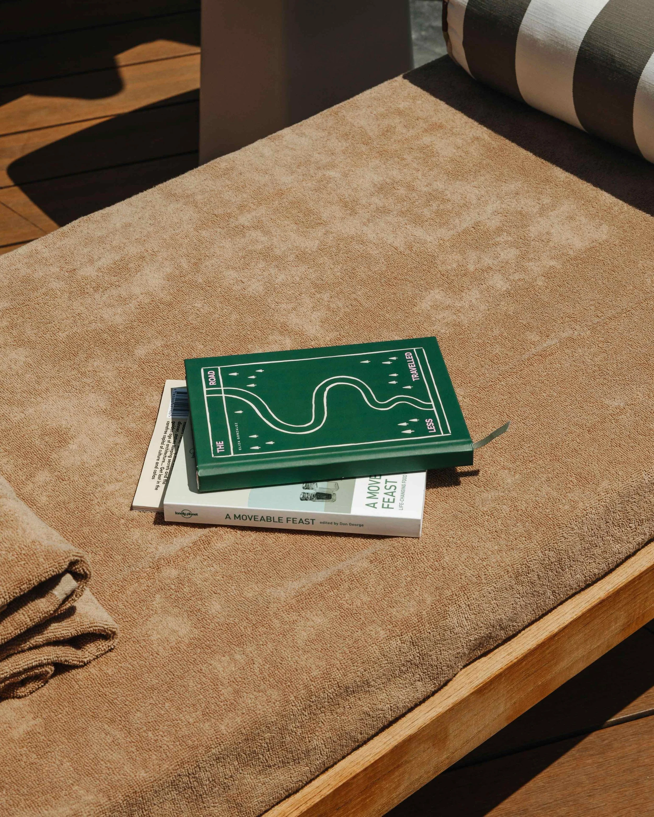 Two books stacked on a beige outdoor table with a folded towel nearby. The top book has a green cover with a map-like design and the bottom book has a white cover titled 'A Moveable Feast'.