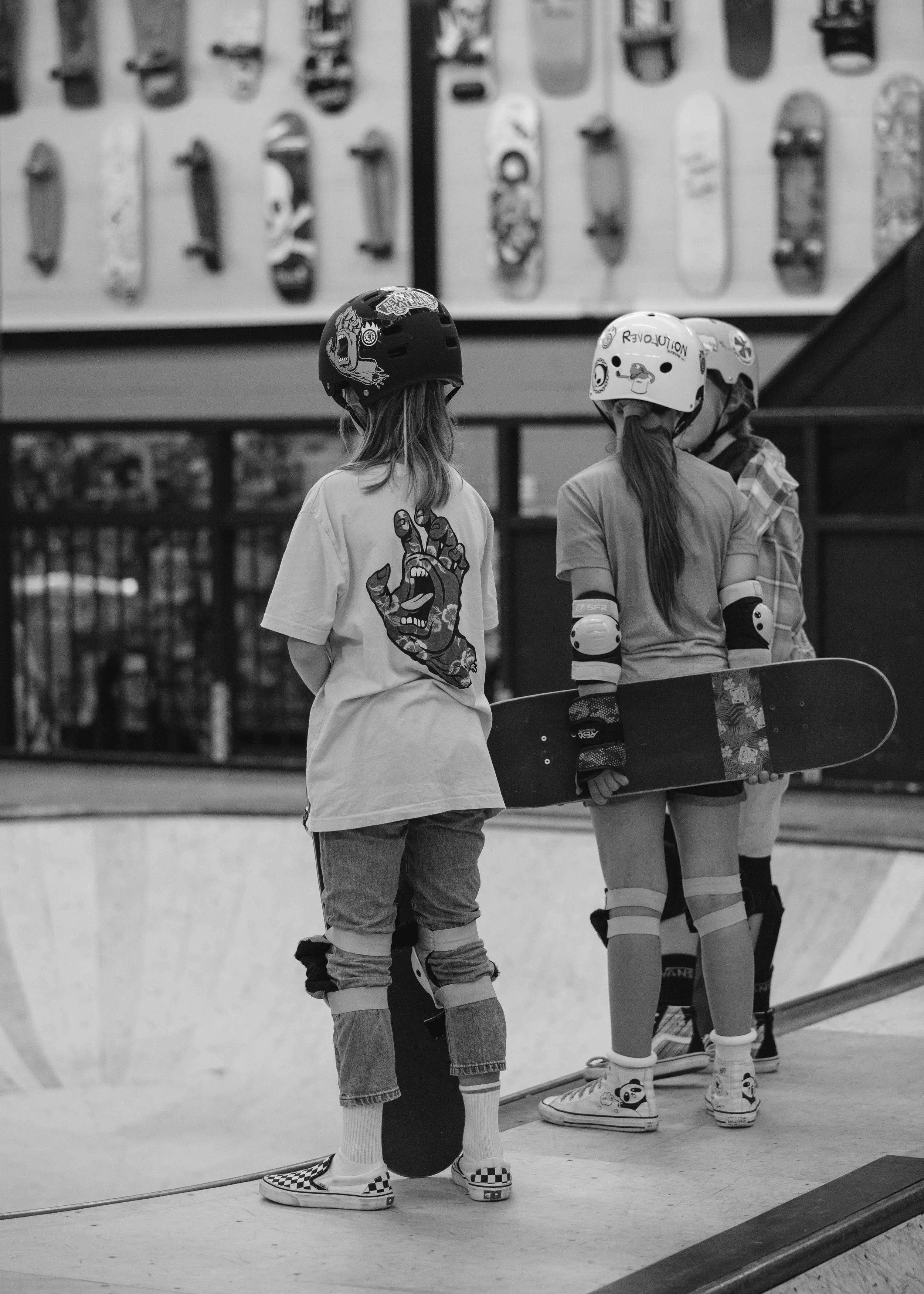 Three young girls at an indoor skate park, two wearing helmets and protective gear, one holding a skateboard, in front of a wall display of skateboards.