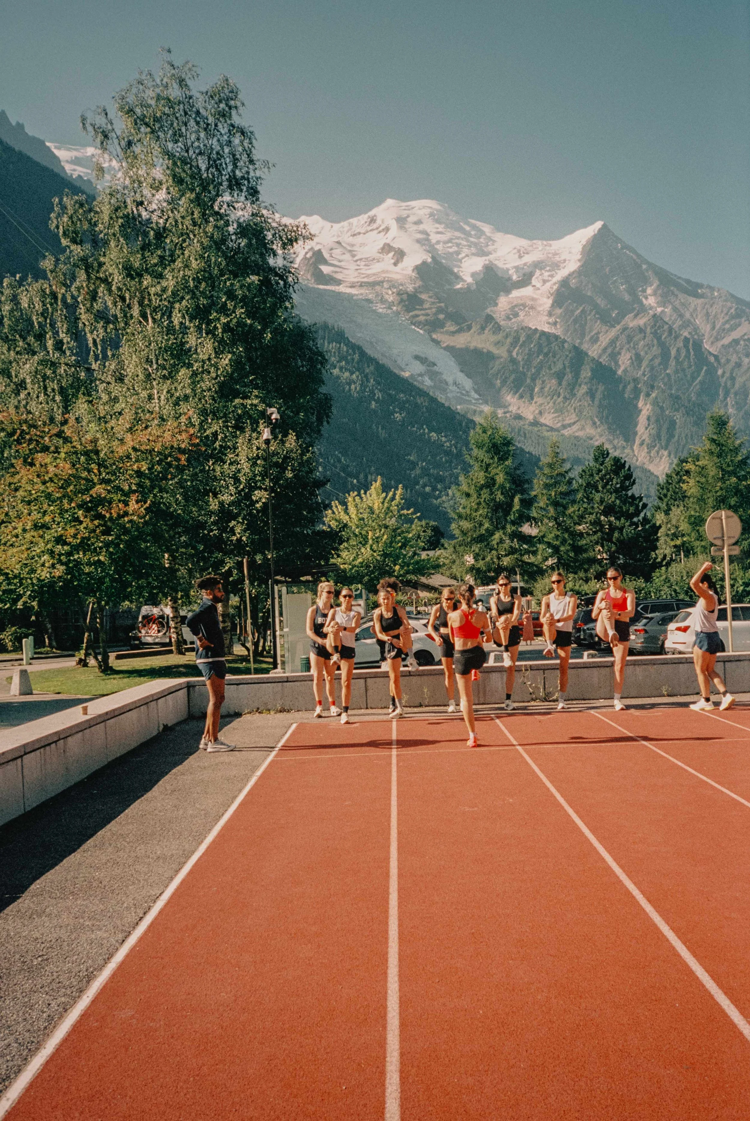A group of young women in athletic clothing stretching on a red running track, with mountains and trees in the background on a sunny day.