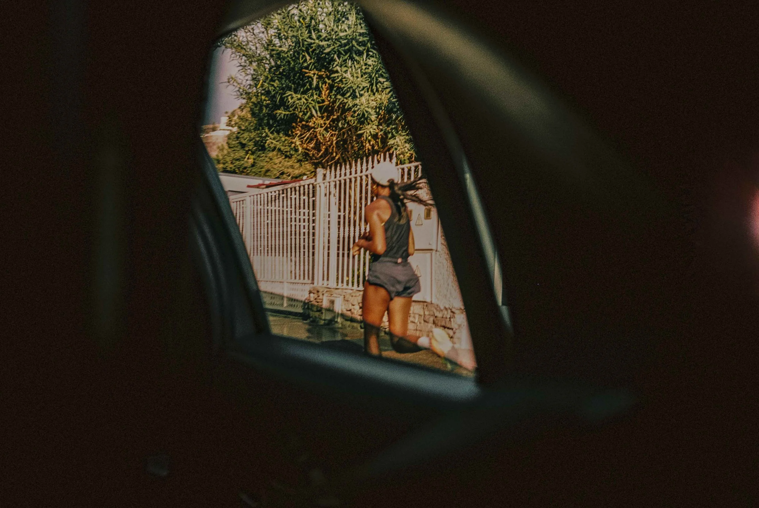 View of a woman jogging outside, seen through a car's side mirror, wearing a black sports outfit and a white cap, next to a fence and a tree.