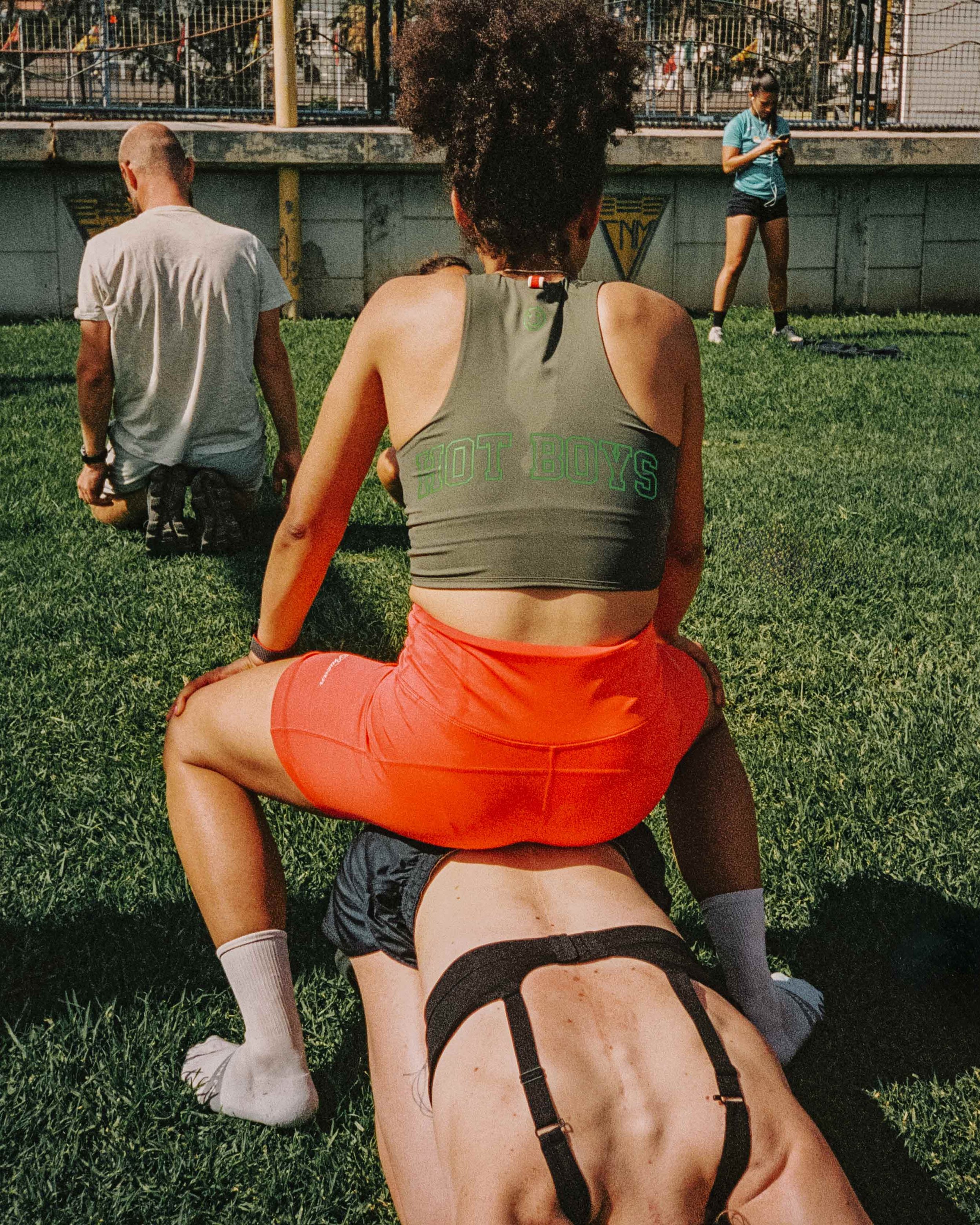 A woman sitting on a man's back on a grassy field, with two other people kneeling and one woman standing in the background at a park or sports field.