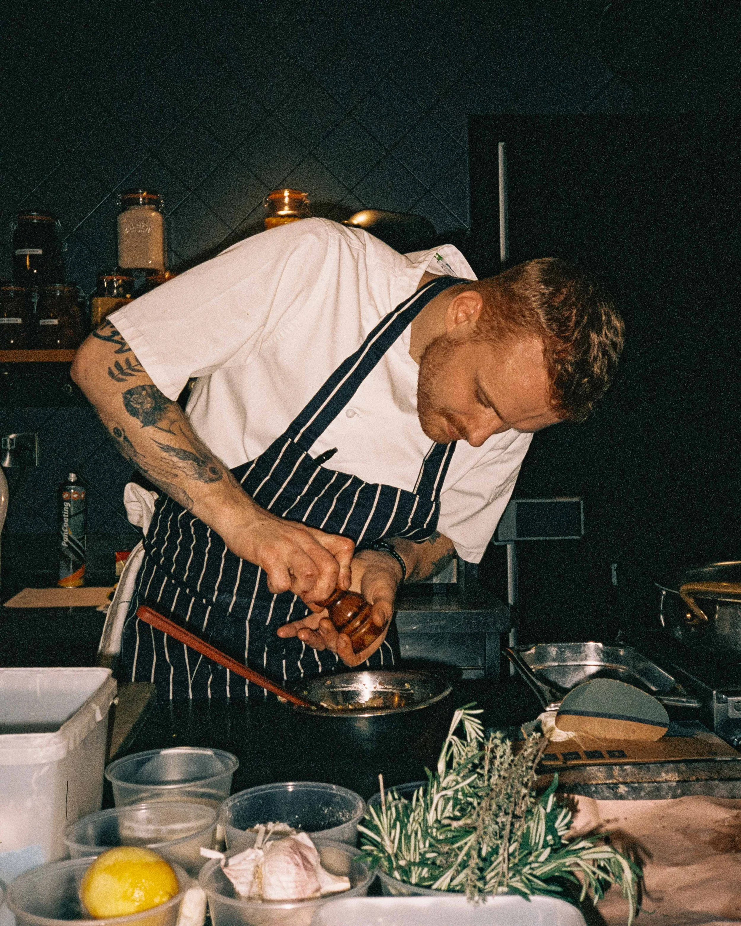 A chef in a striped apron preparing food in a professional kitchen, surrounded by ingredients and utensils.