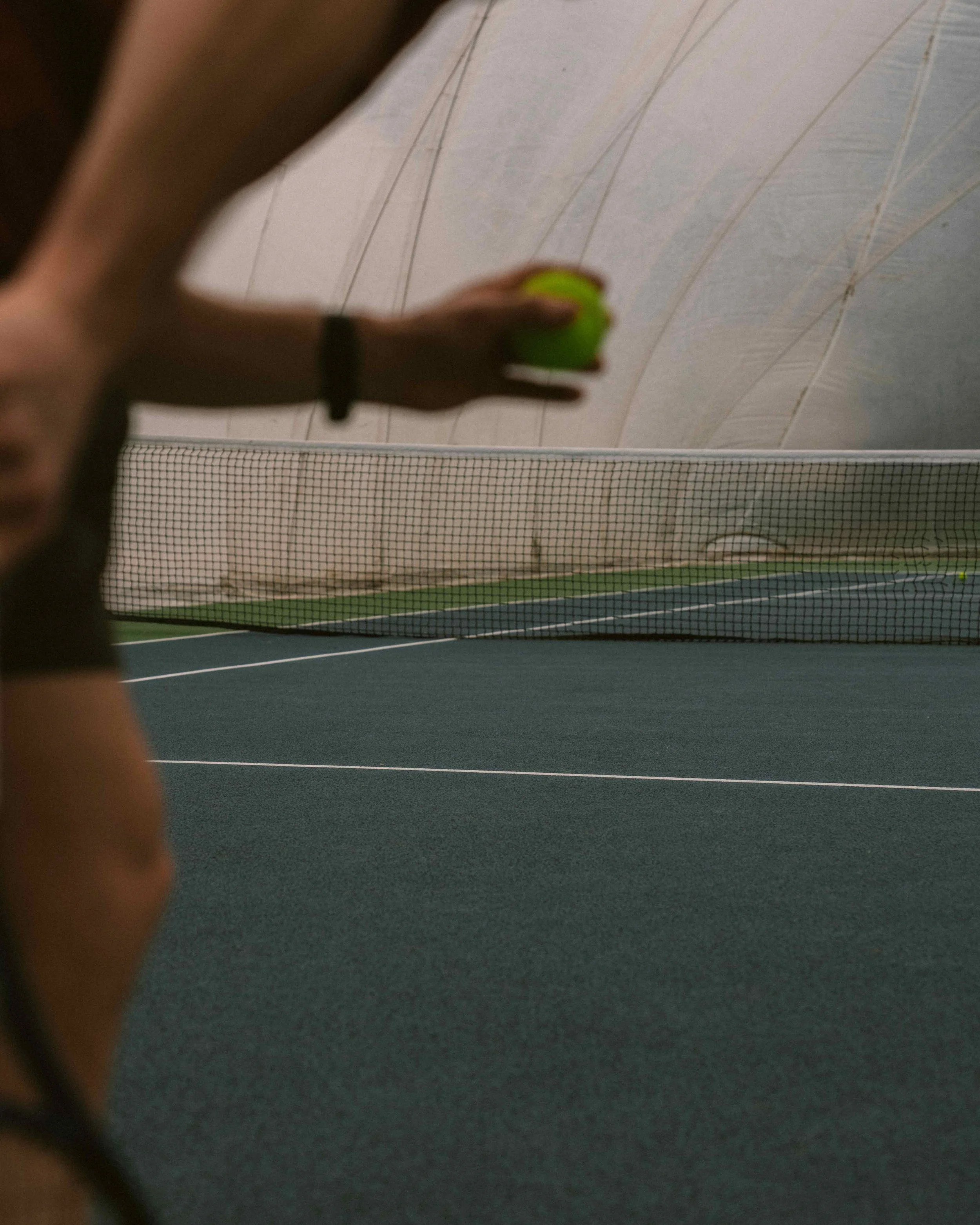 Person holding a tennis ball on an indoor tennis court with a net in the background.