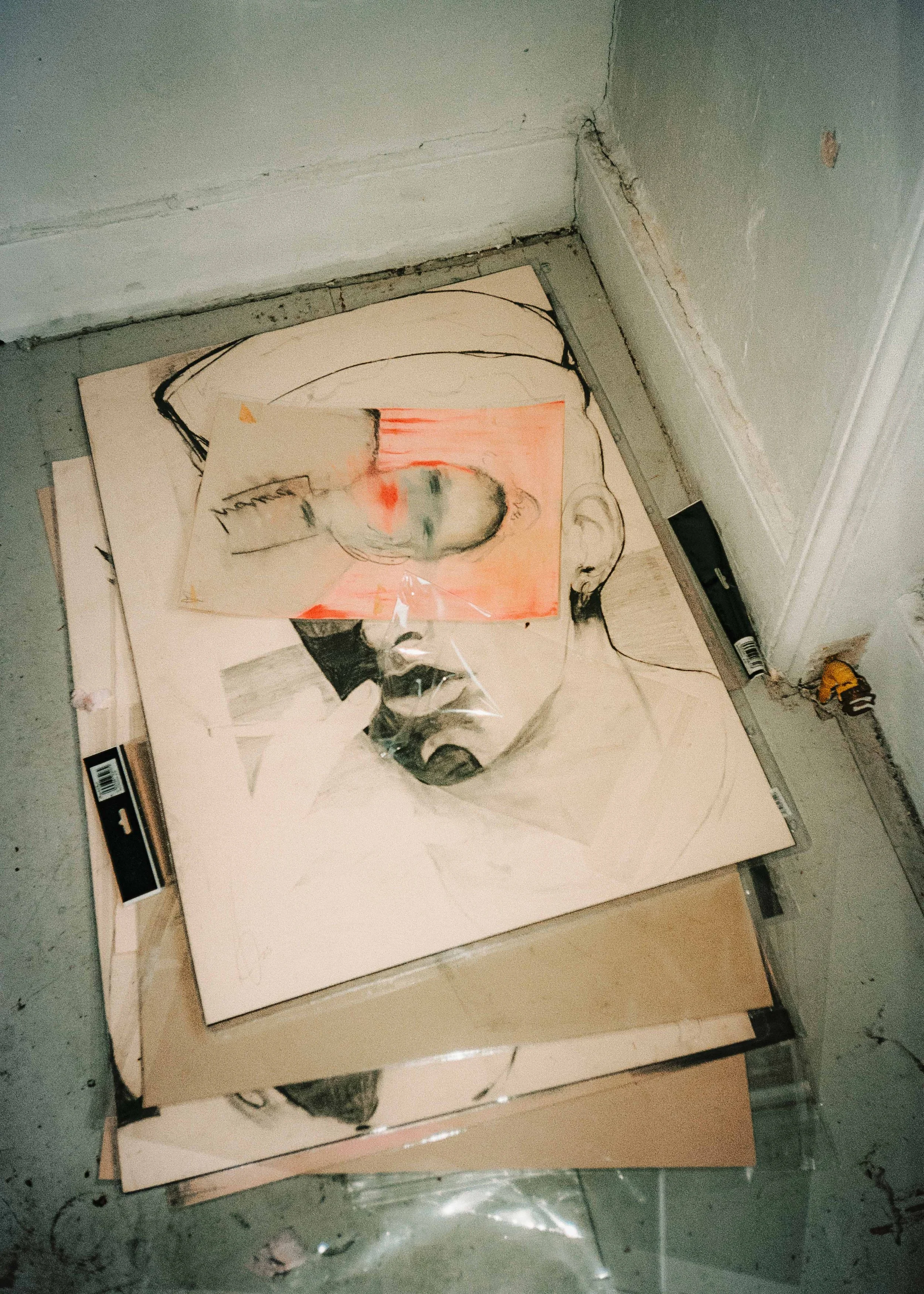 Stack of drawings on the floor, including a portrait sketch of a person with glasses and a small abstract watercolor face overlaying the sketch.