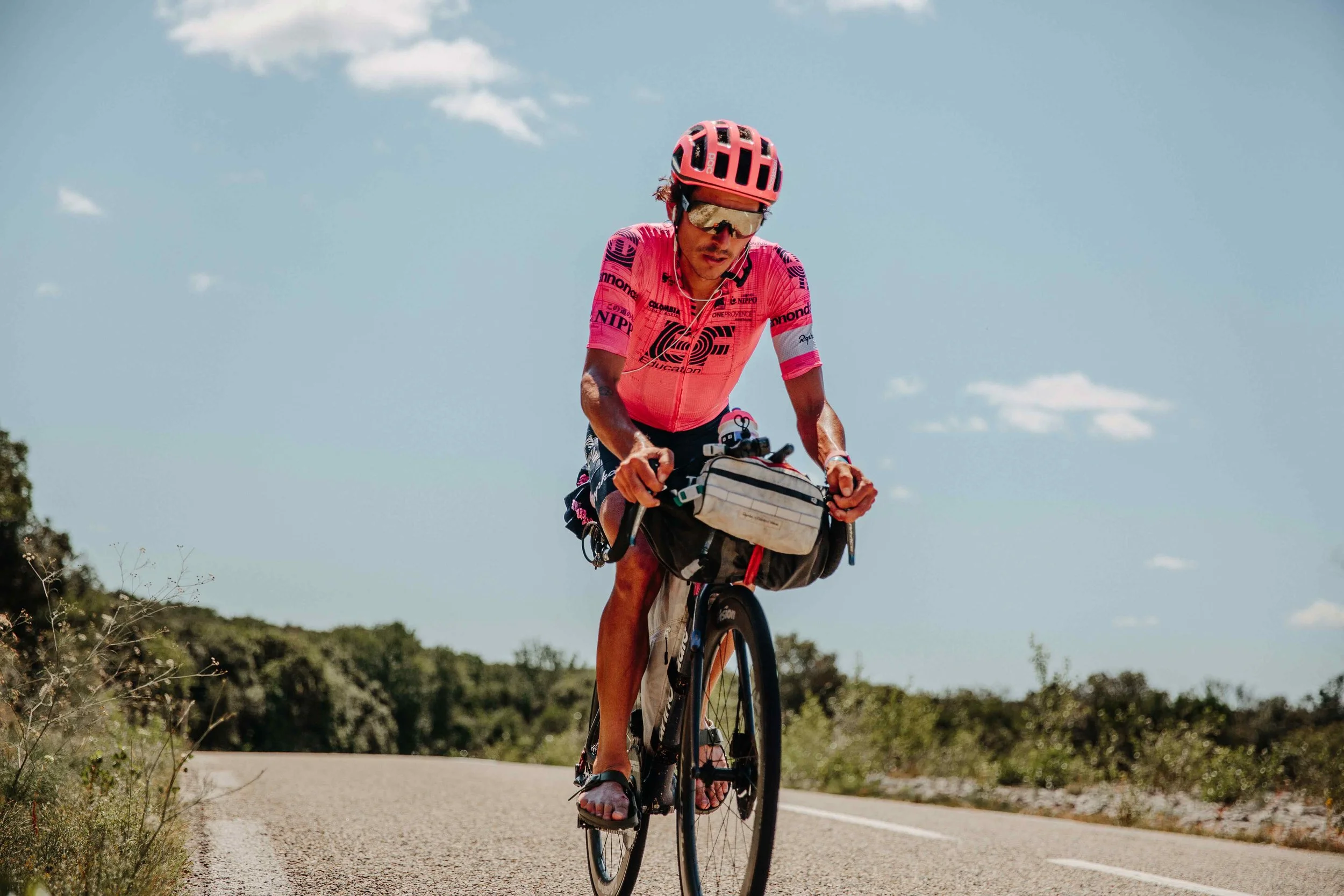 A person riding a bicycle on a rural road, wearing a pink cycling jersey, a pink helmet, and sunglasses, with a small bag attached to the bike.