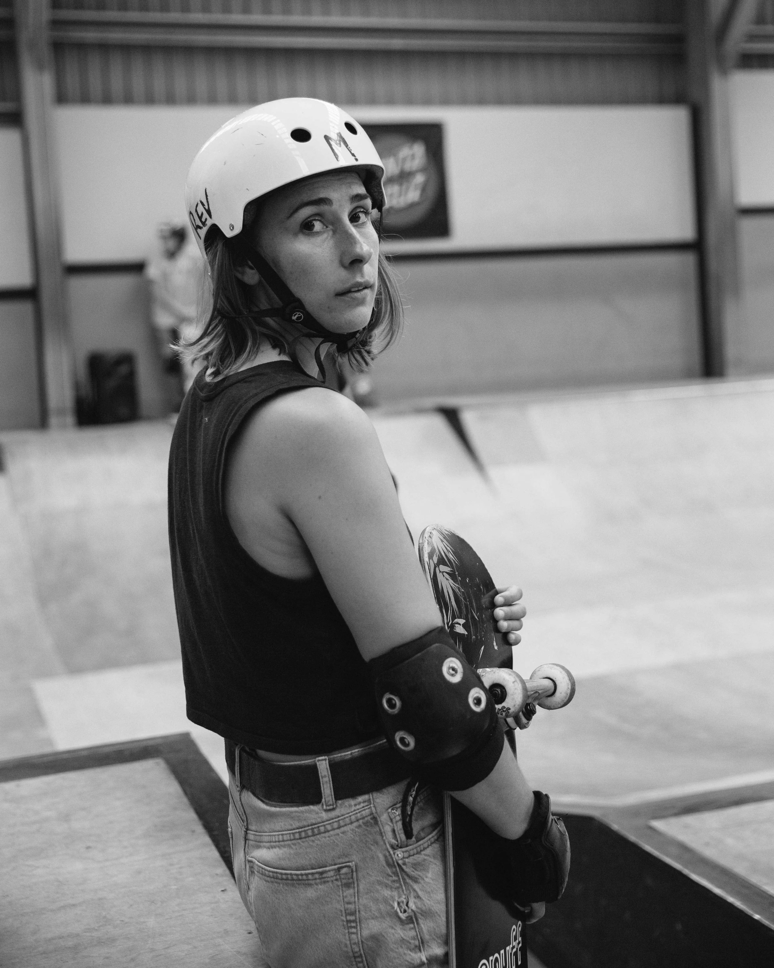 A young woman wearing a helmet, elbow pads, and sleeveless shirt holding a skateboard at an indoor skate park.