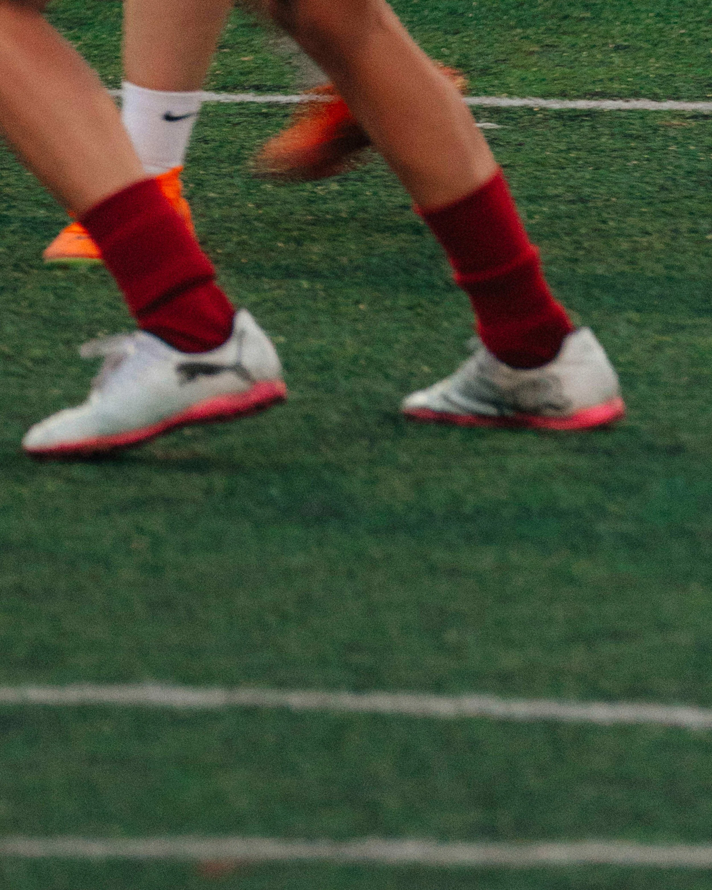 Close-up of a soccer player's legs and feet on an artificial turf field, wearing white cleats, red socks, and orange cleats visible in the background.