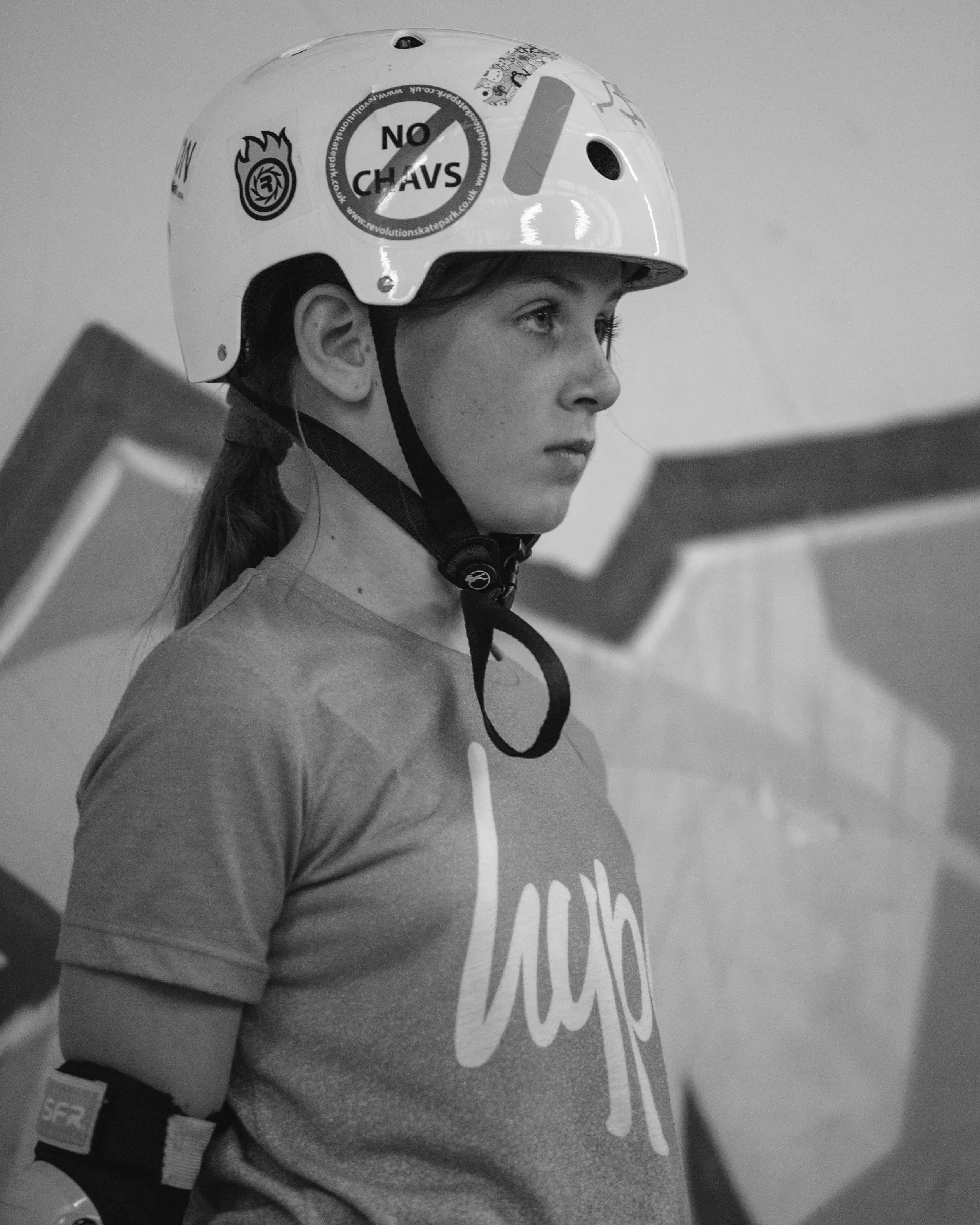 A young woman wearing a helmet and a T-shirt stands indoors with a serious expression, possibly at a sports or climbing facility.
