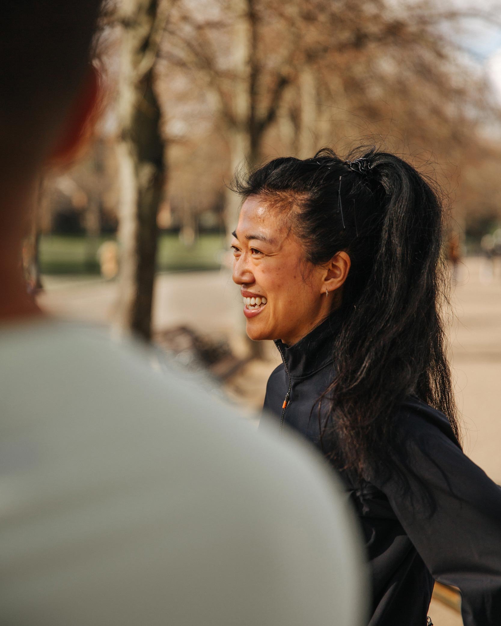 A woman with long black hair, smiling and talking outdoors in a park with trees in the background.
