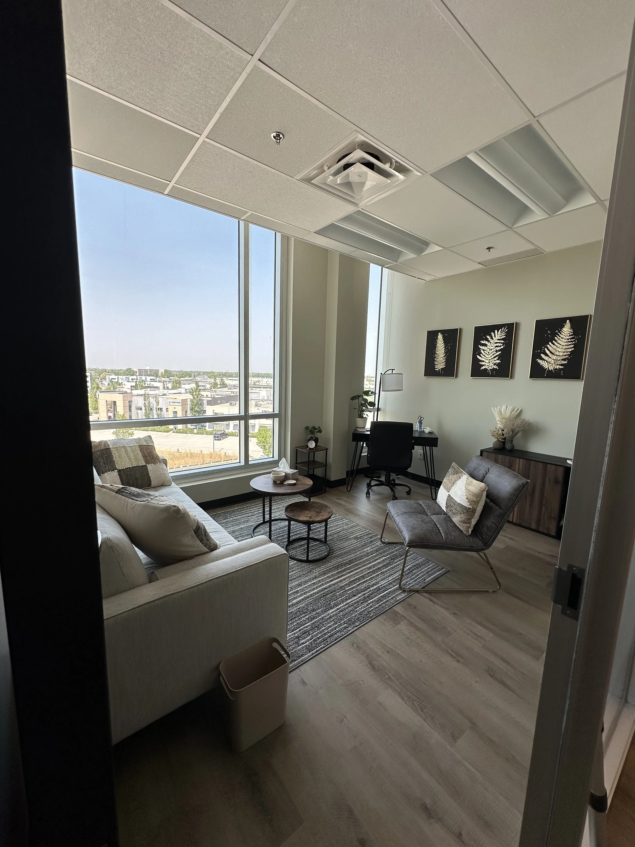 Modern office room with large window, beige sofa with pillows, black office chair, small round tables, grey chair, framed fern art on wall, desk with lamp, decorative plants, and light wooden flooring.