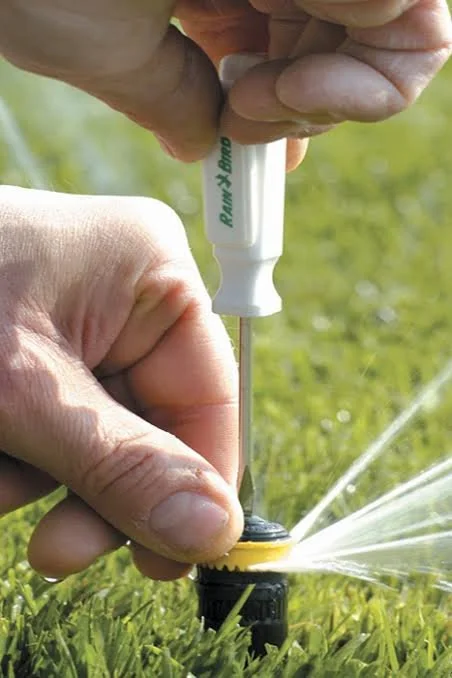 Close-up of a person's hand adjusting a sprinkler head in a grass lawn with water spraying out.