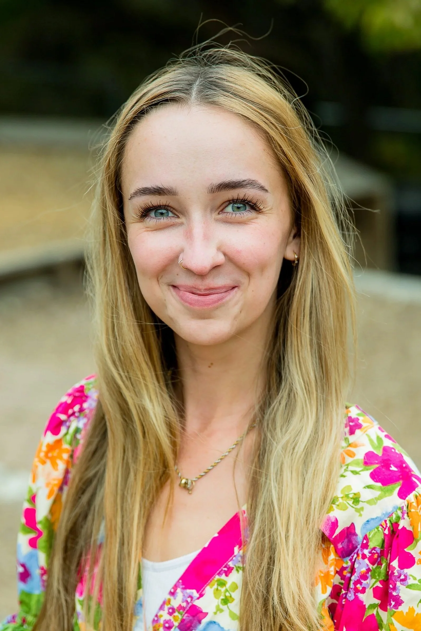 A young woman with long blonde hair and bright blue eyes, smiling outdoors with a blurred natural background.