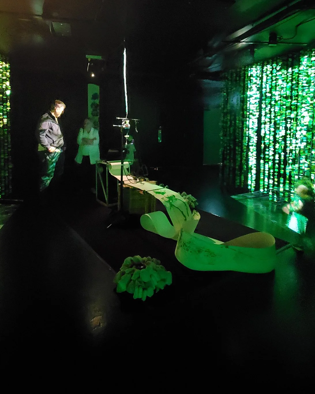 Photo documentation of Arboreal Intuitions. Two people stand next to a custom made drawing plotter. A long roll of paper is fed through the plotter platform and drawings of tree growth rings are made.