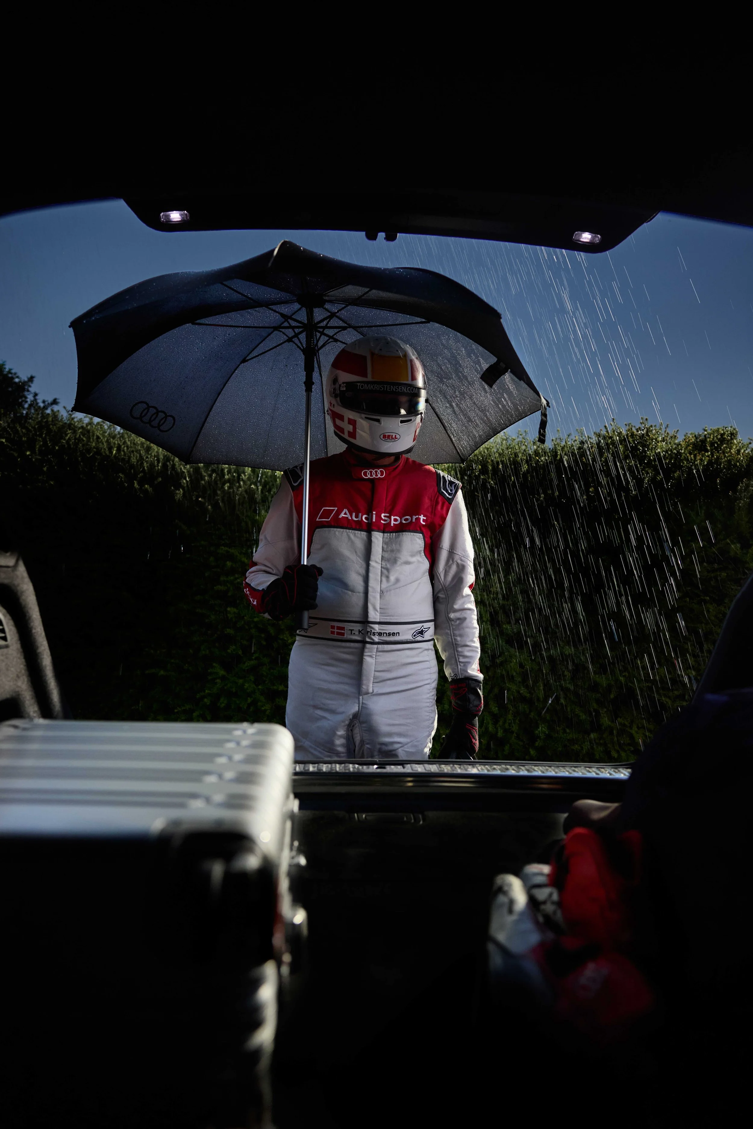 Tom Kristensen in full race suit and helmet holding an umbrella in rainy weather seen through the open trunk of an Audi RS Q8 performance at Goodwood Festival of Speed