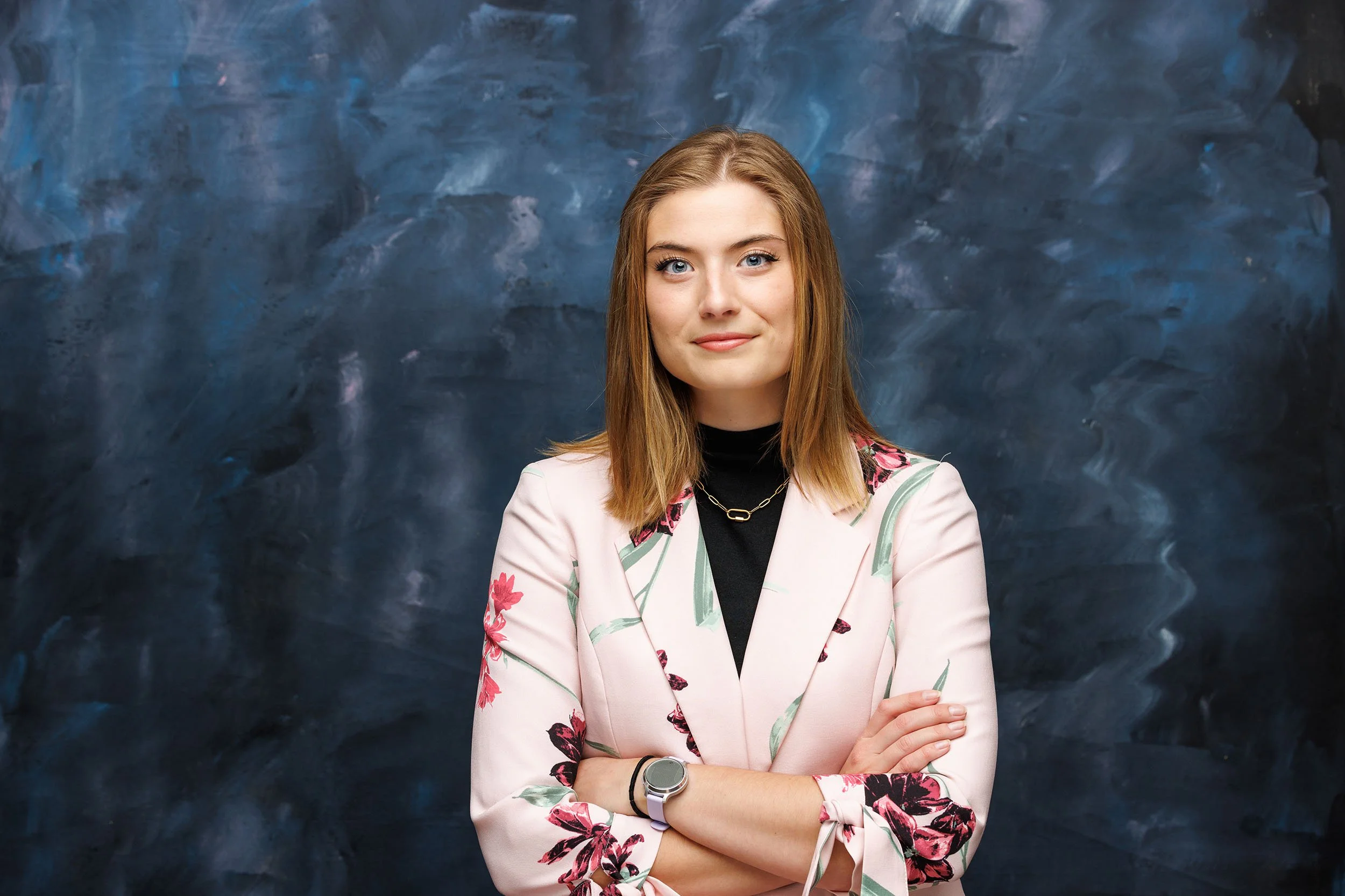 Woman's headshot: wearing a pink blazer with flowers, against a dark blue painted background her arms are folded